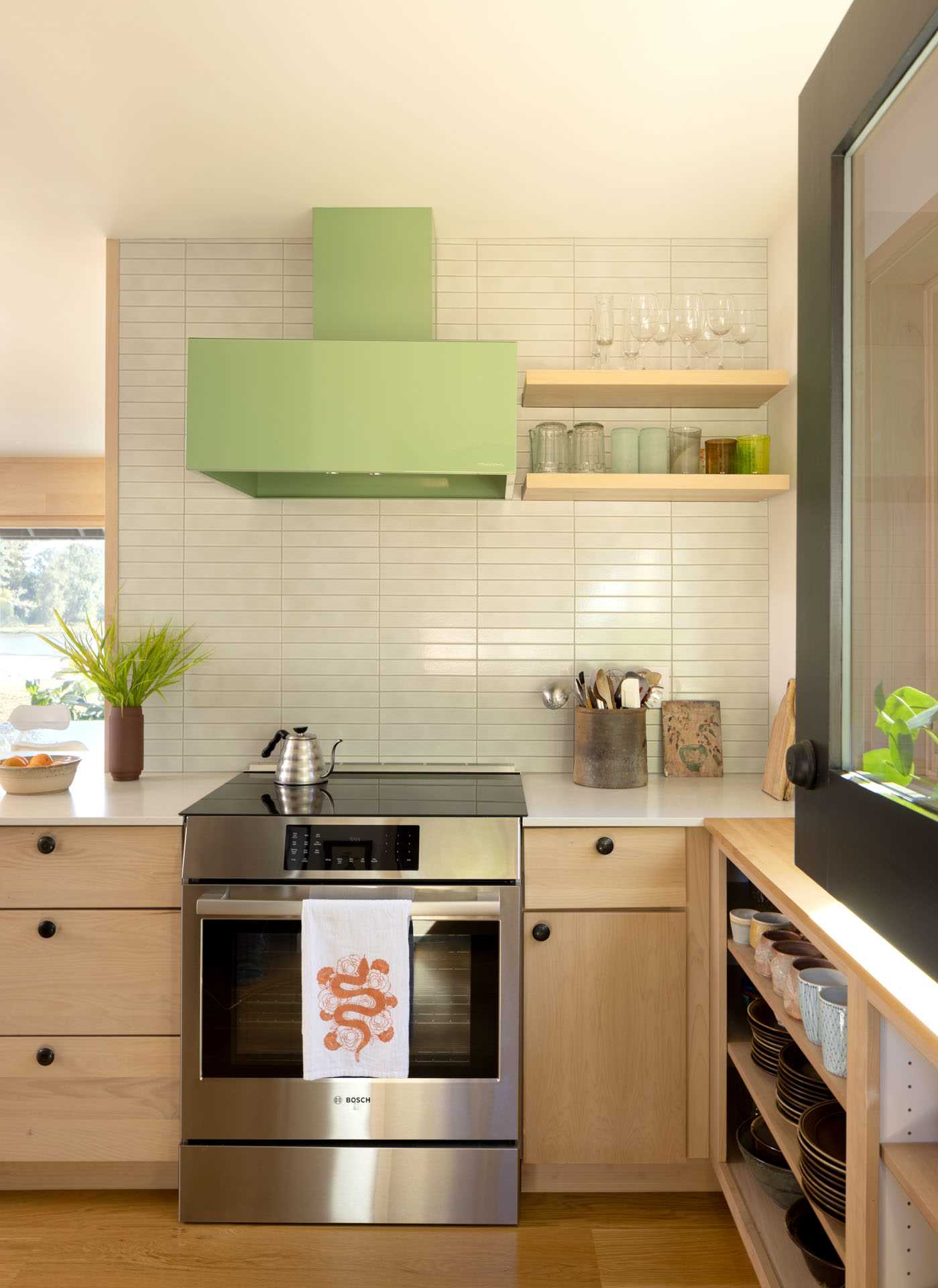 آشپزخانه بازسازی‌شده با کابینت‌های چوبی سبک و قفسه‌های باز، هود سبز رنگ پاستلی، دیواری با کاشی‌های روشن، و کف‌های چوبی.  یک درب هلندی نیز اضافه شد.