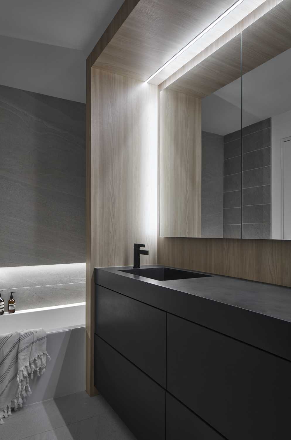 A modern bathroom with a dark vanity that has a wood surround with embedded lighting, and a built-in bathtub.