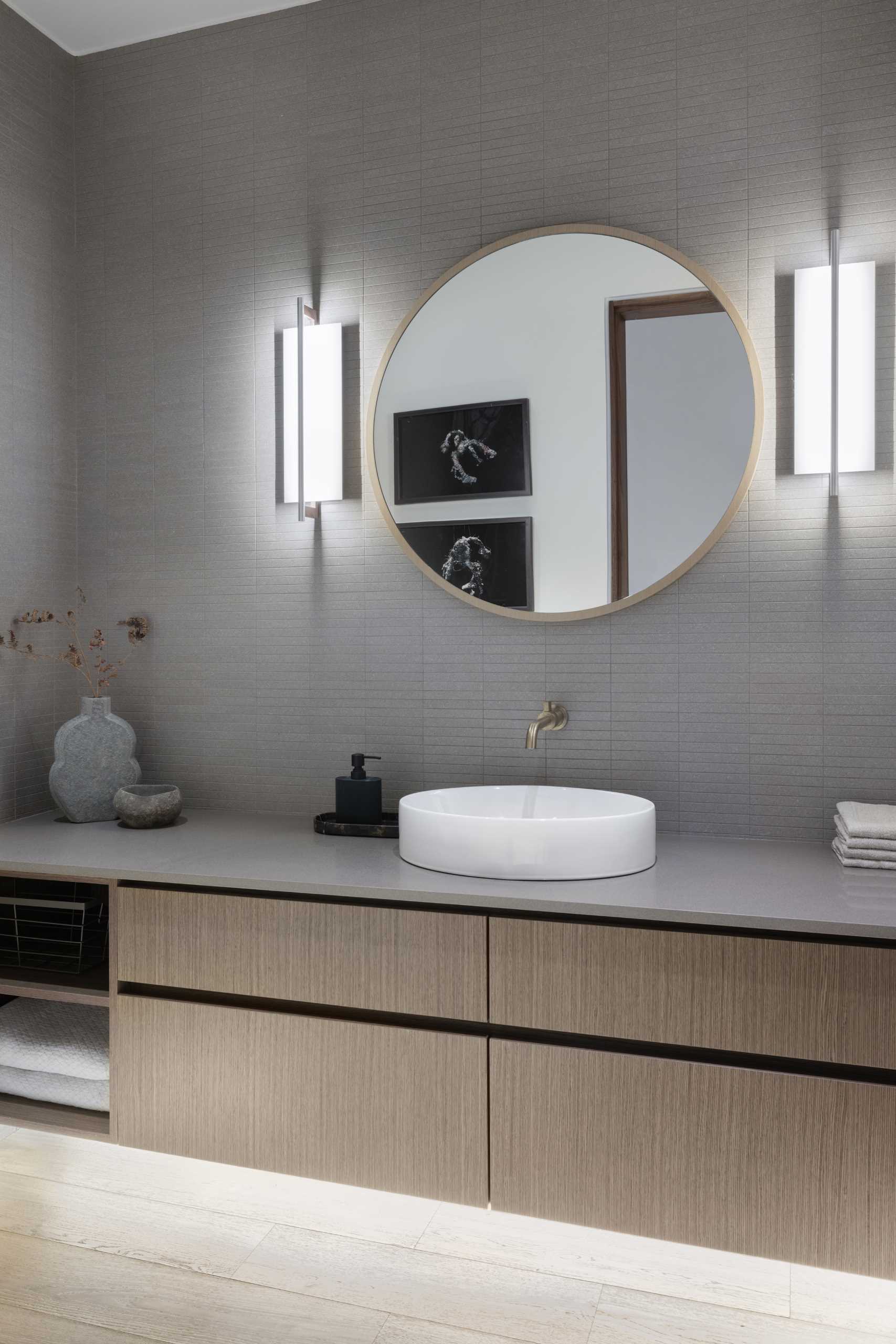 A modern bathroom with grey tiles, a wood vanity, and round mirror.