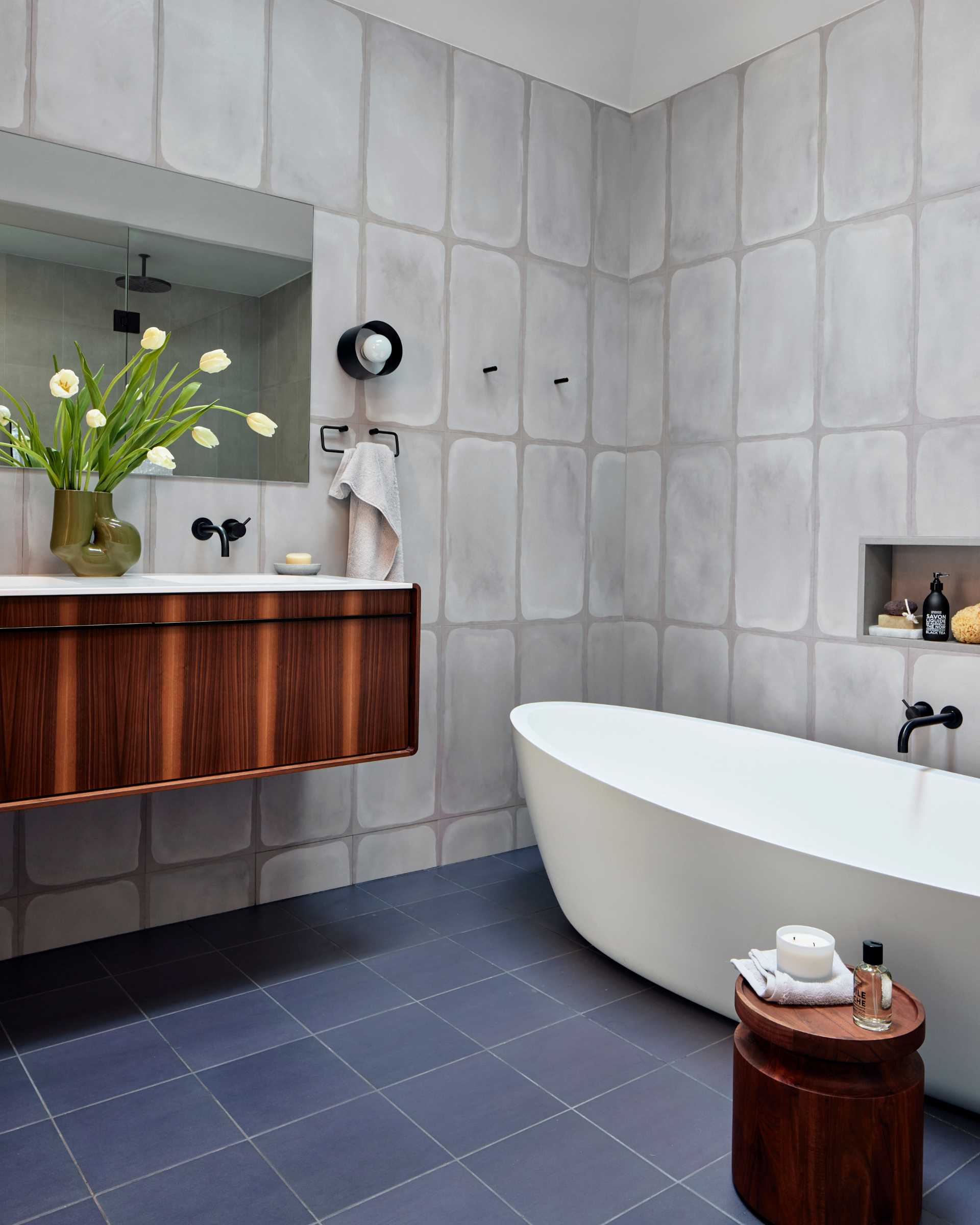 A modern bathroom with a vaulted ceiling, tile-covered walls and floor, a freestanding bathtub with a shelving niche, and a wood vanity.