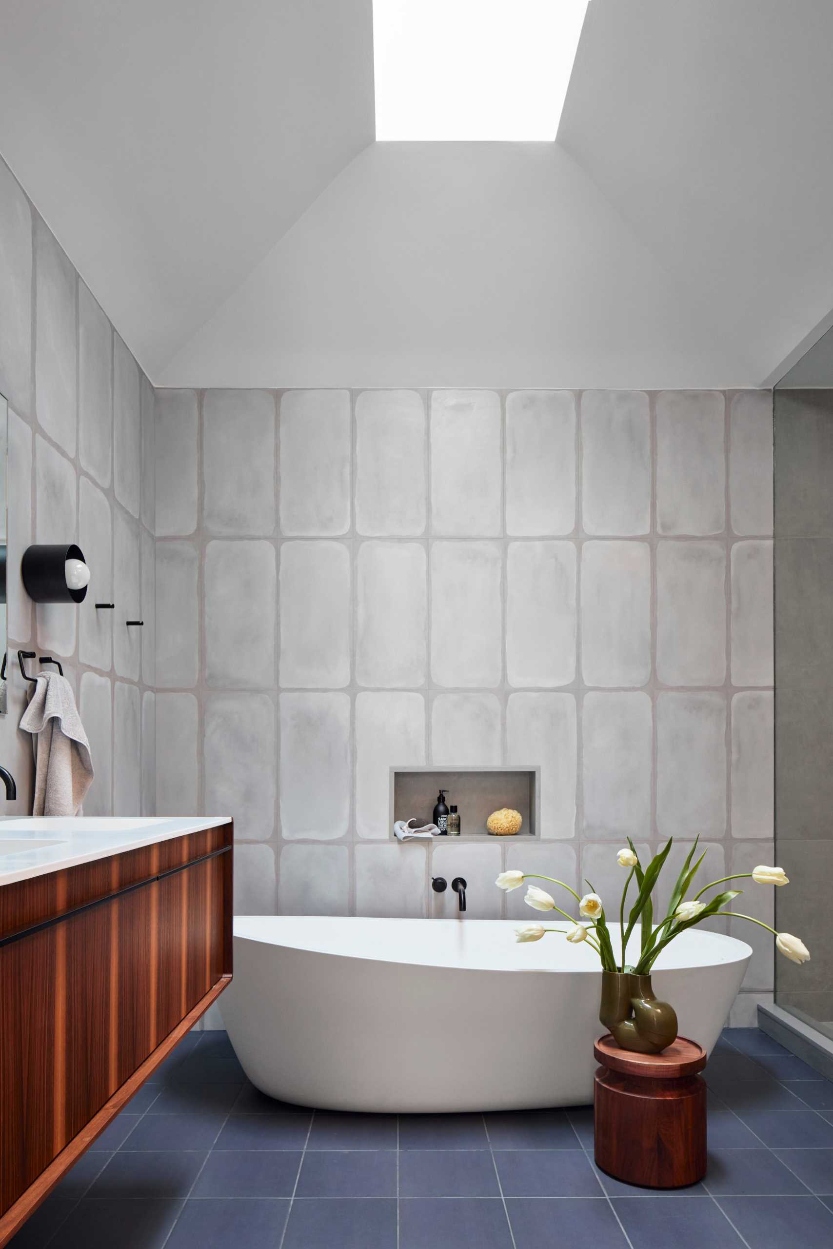 A modern bathroom with a vaulted ceiling, tile-covered walls and floor, a freestanding bathtub with a shelving niche, and a wood vanity.