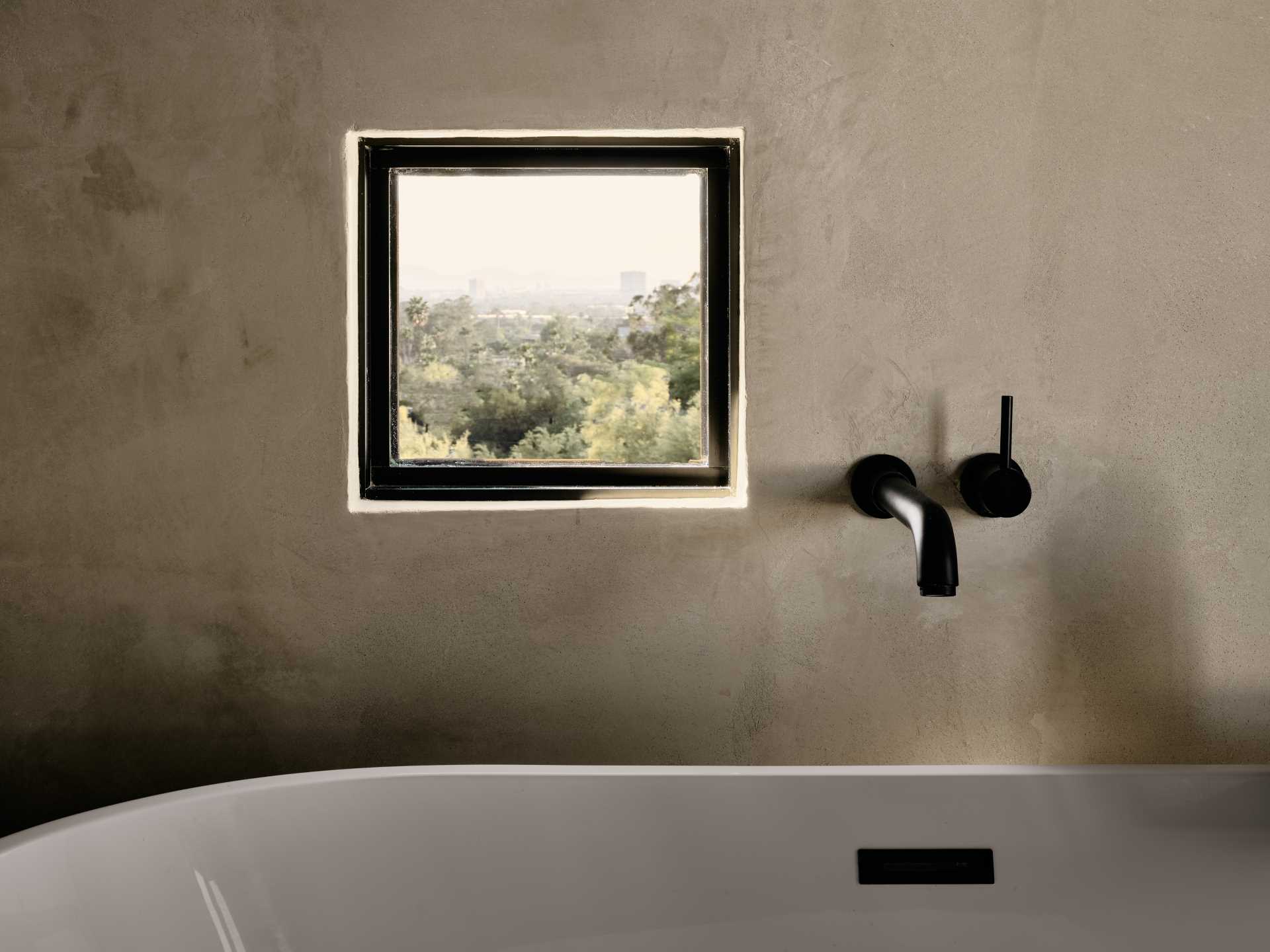 A modern earthy bathroom with a freestanding bathtub and a small square window.