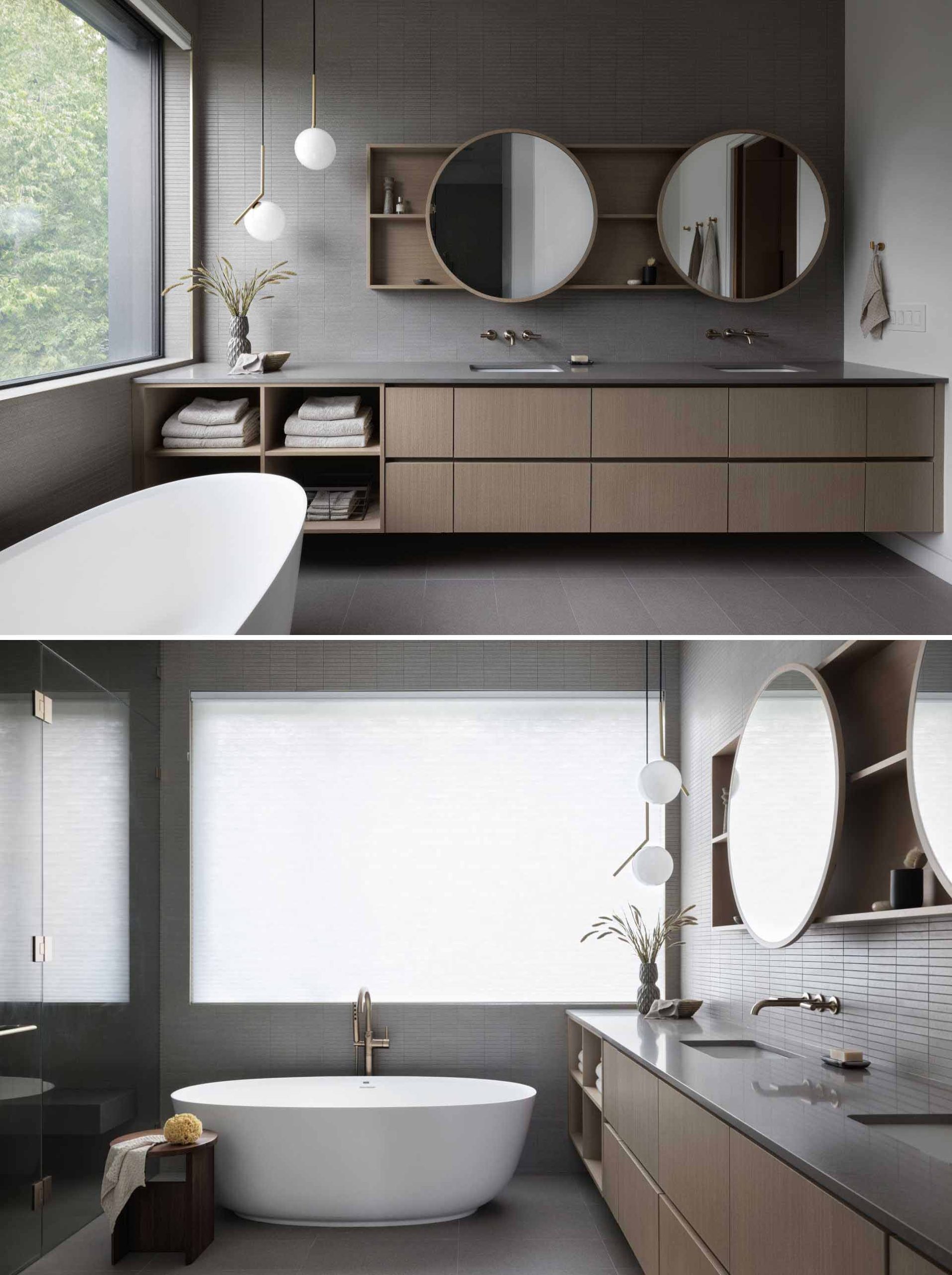 A modern bathroom with grey tiles, a wood vanity, round mirrors, freestanding bathtub, and glass-enclosed shower.