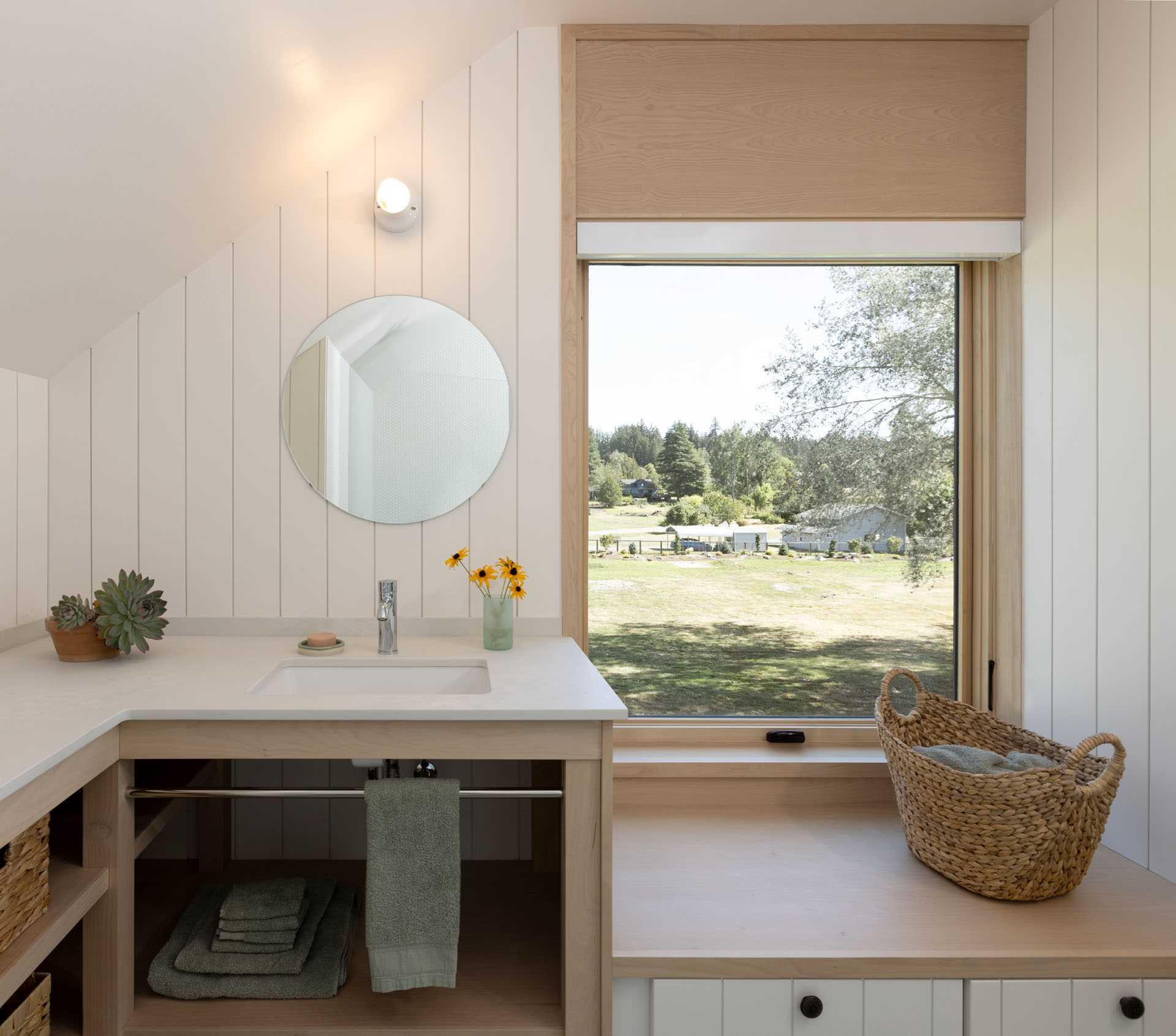 A modern bathroom with white walls, a round mirror, and a bench under the window.