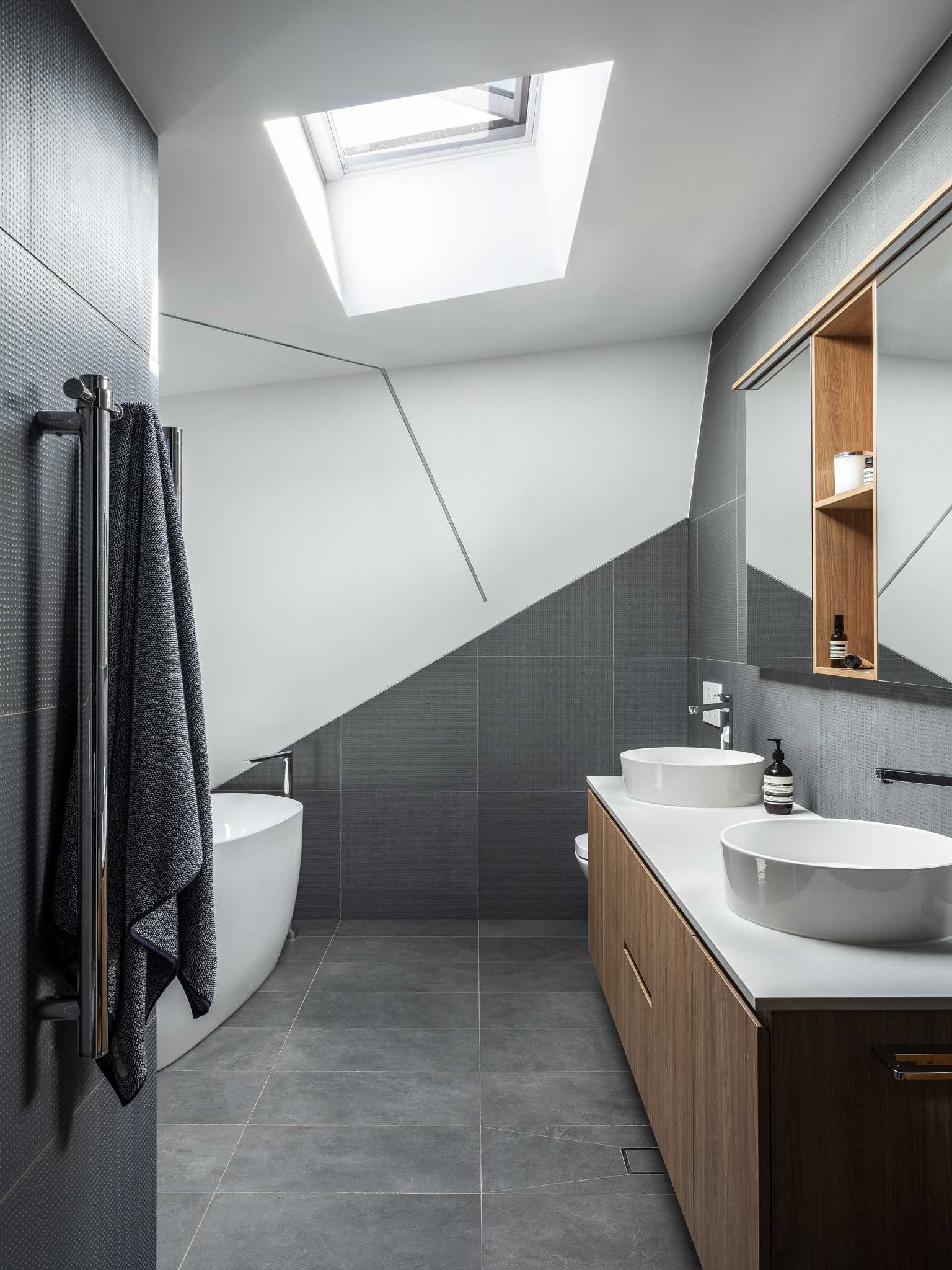 In this bathroom, grey tiles partially installed with a diagonal design add visual interest and are paired with white walls and a wood vanity. A skylight helps to keep the bathroom bright.