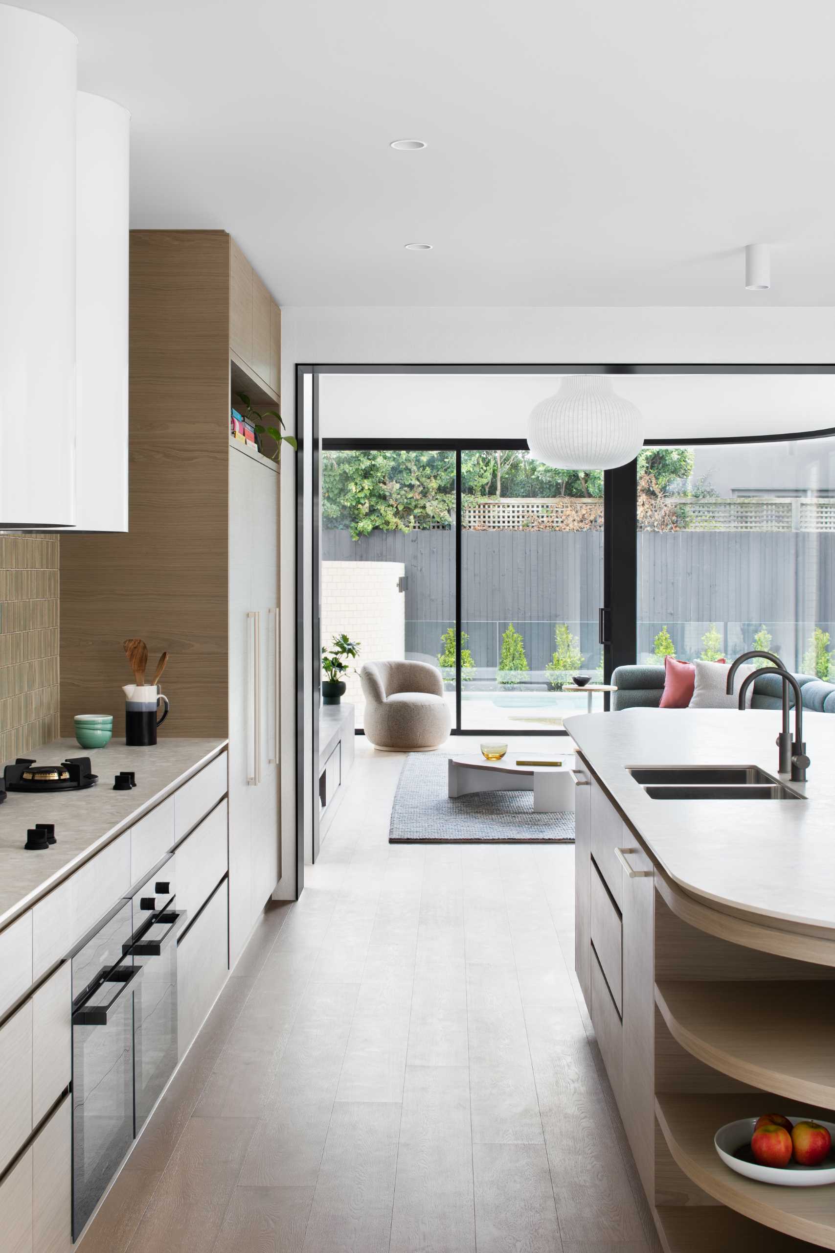 This modern kitchen features a tiled wall, wood cabinets, and an island with curved ends. One end is dedicated to seating, while the other has open shelving.