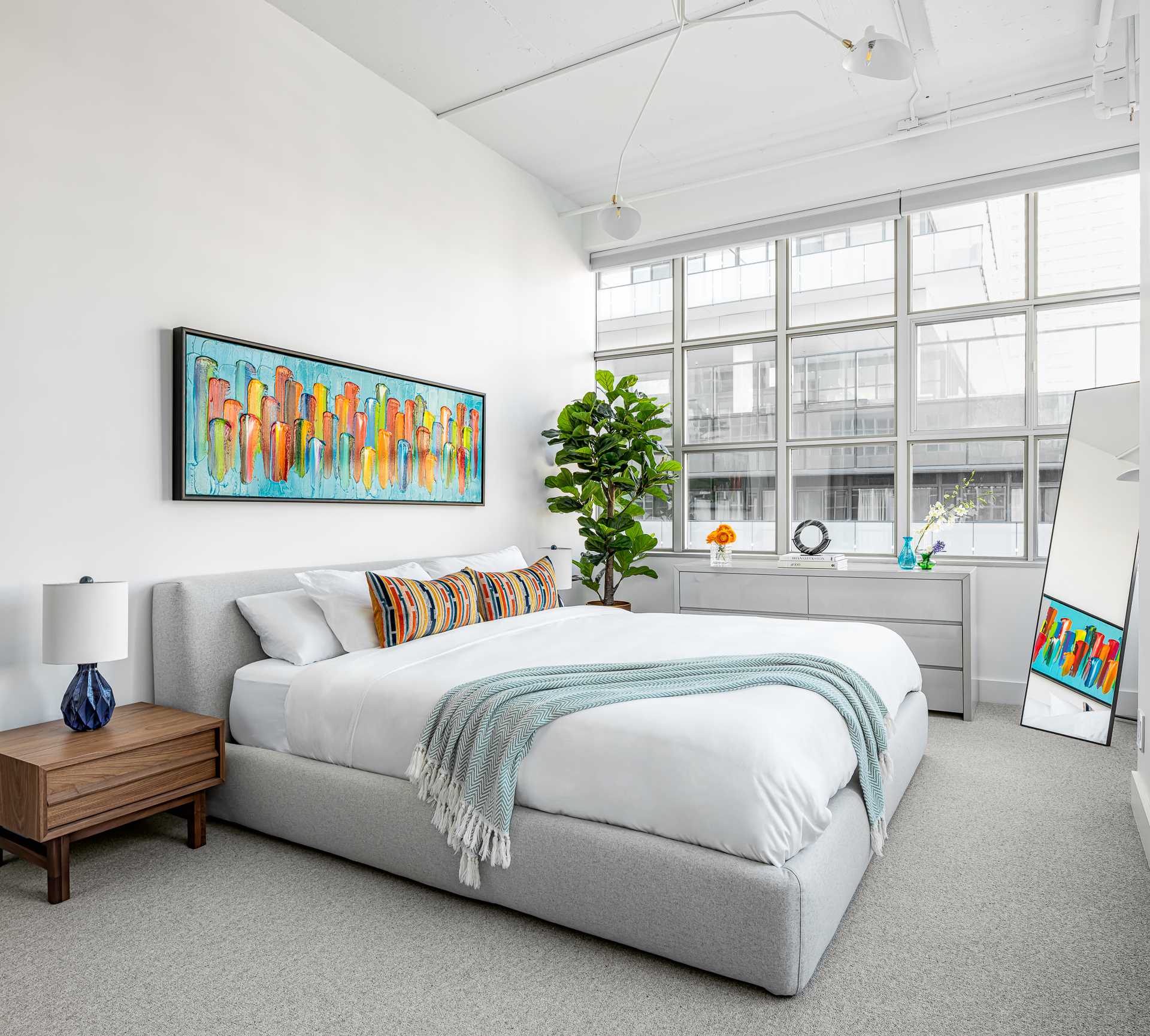 This contemporary primary bedroom in a loft apartment has a subtle color palette, with a large king bed in a soft grey wool fabric. 