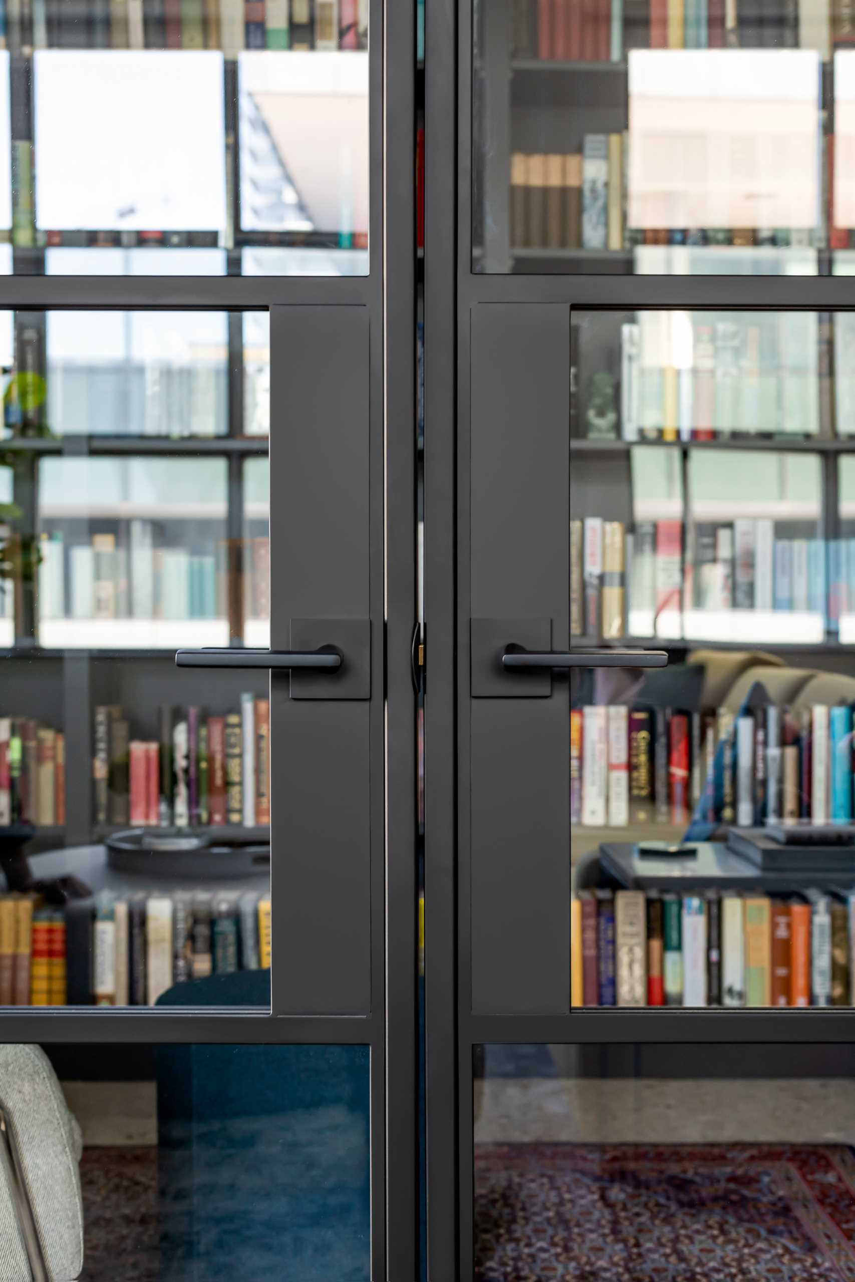 A modern home library enclosed behind a glass wall, has custom black shelving and a ladder to reach the upper shelves.