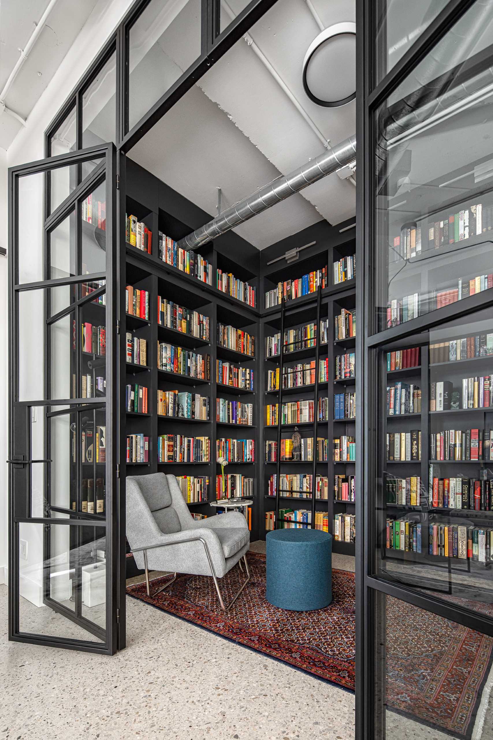 A modern home library enclosed behind a glass wall, has custom black shelving and a ladder to reach the upper shelves.