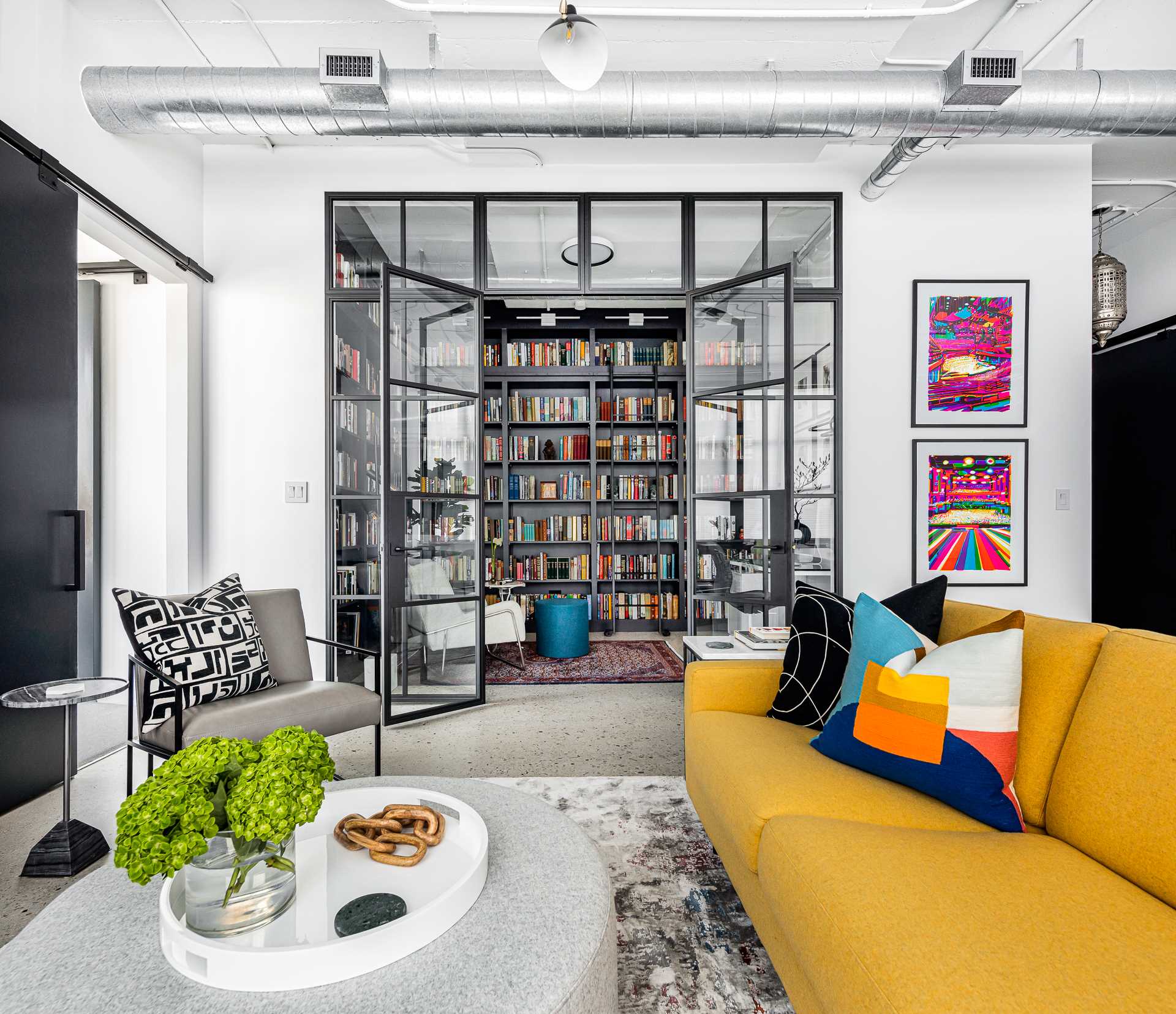 A modern home library in a loft apartment, is enclosed behind a glass wall, has custom black shelving and a ladder to reach the upper shelves.