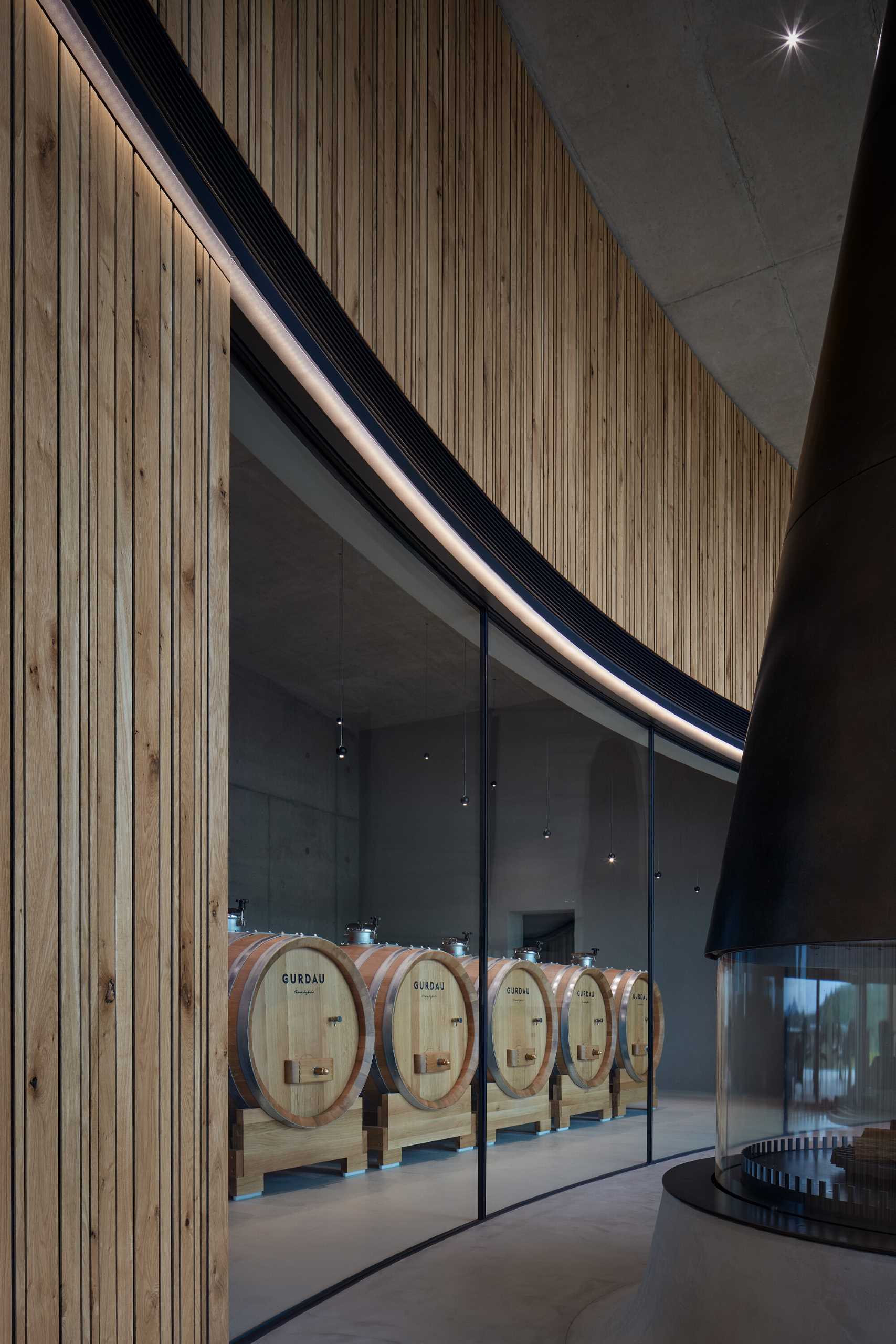 مجموعه‌ای از بشکه‌های شراب در پشت یک دیوار شیشه‌ای منحنی قرار گرفته‌اند و به بازدیدکنندگان نگاهی اجمالی از فرآیند تولید شراب می‌دهند.