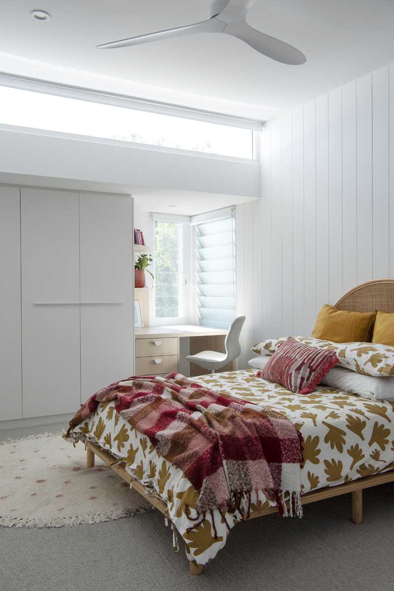 A modern bedroom has a desk built into the corner by the windows, creating a small study area.