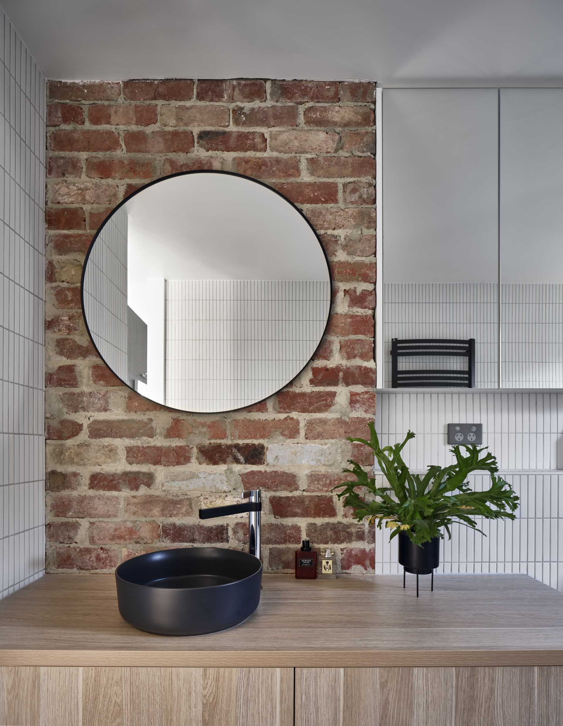 In this modern bathroom, a small brick accent wall provides a backdrop for the round mirror above the vanity, while vertical rectangular tiles cover the other walls.