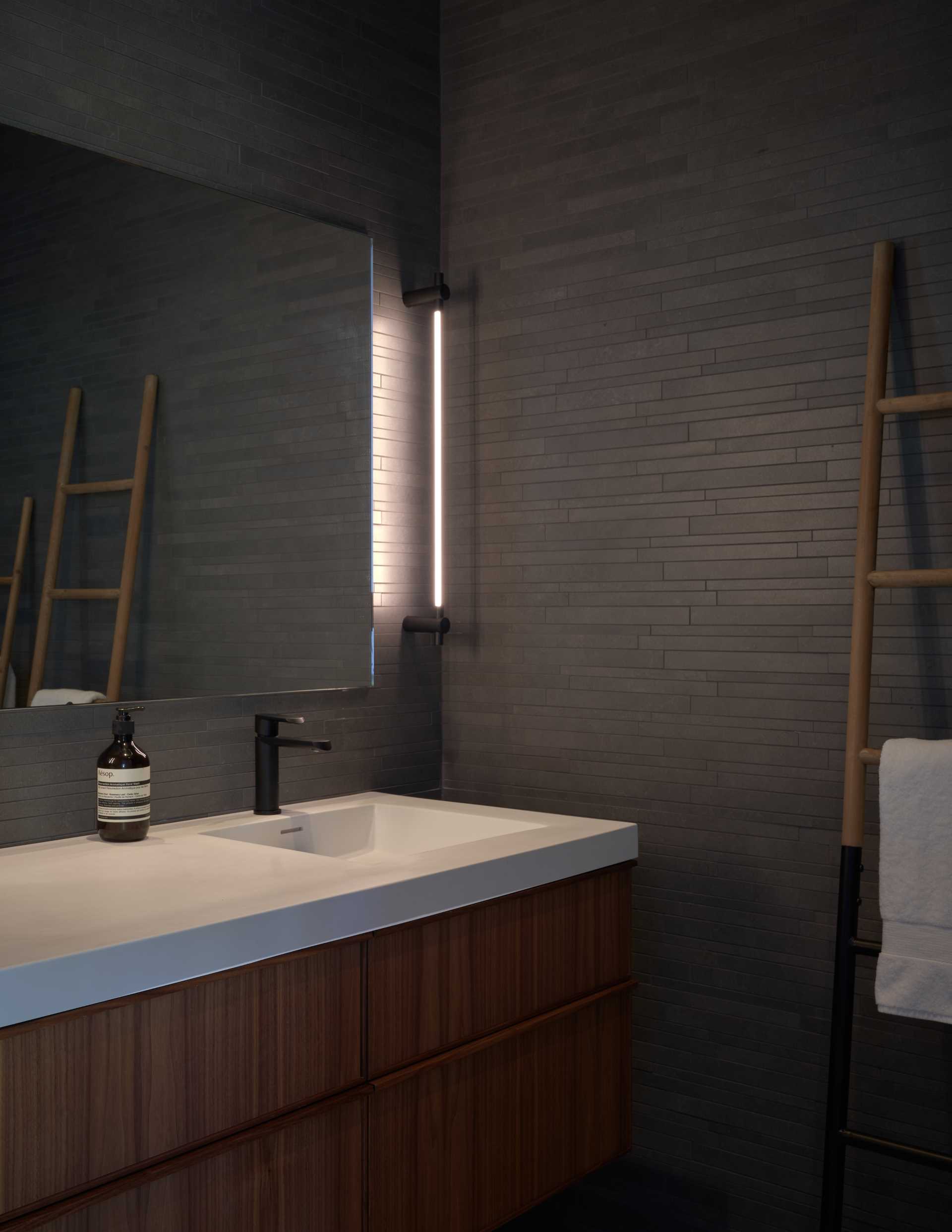 In this modern bathroom, dark tiled walls have been paired with a wood vanity, a light countertop, and vertical lighting.