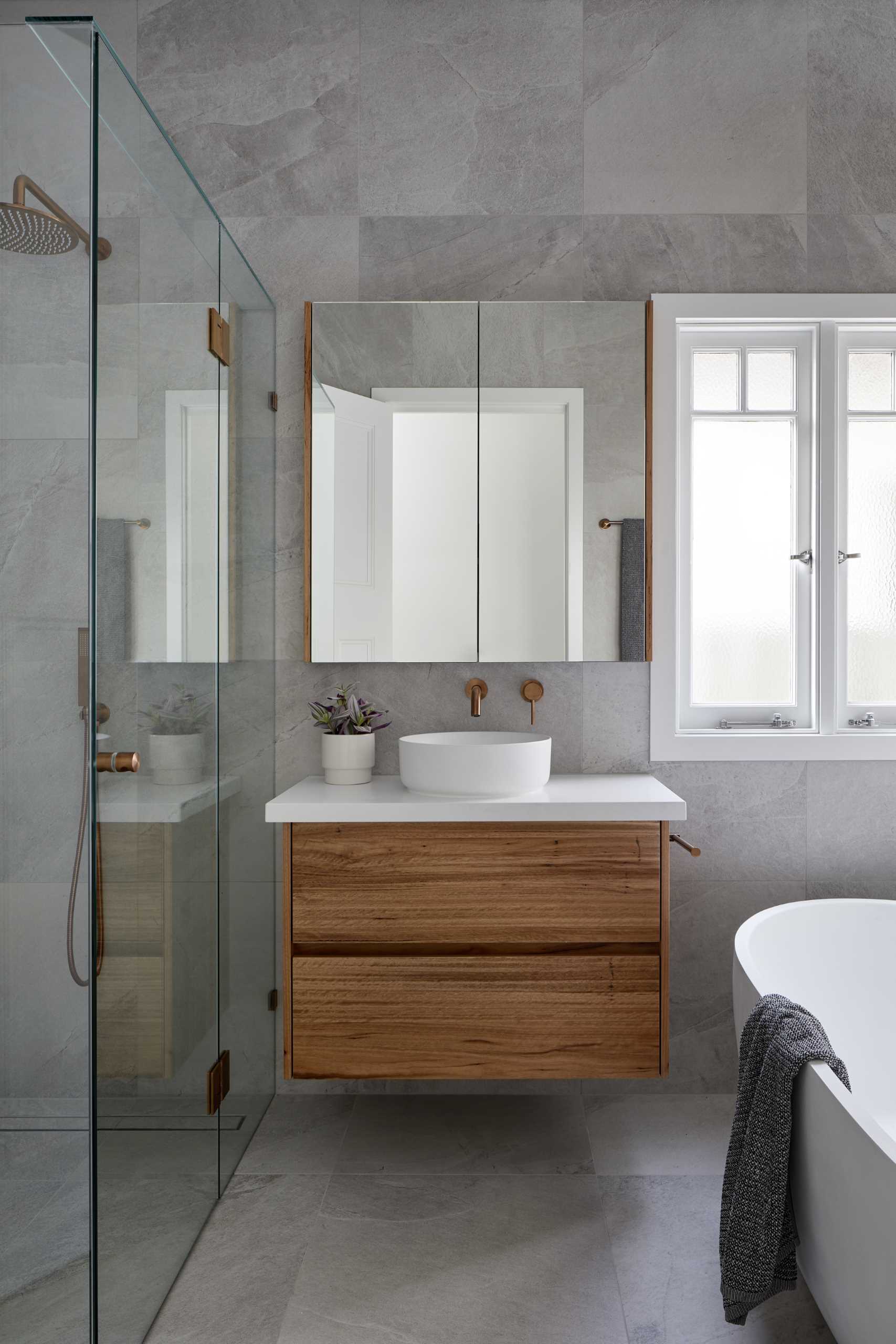 This modern en-suite bathroom has a single vanity with a glass-enclosed walk-in shower on one side and a freestanding bathtub with a shelving niche on the other.
