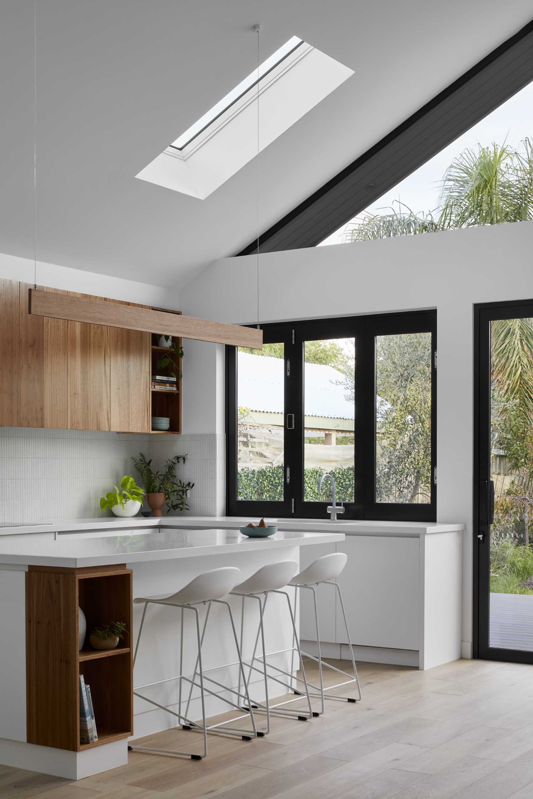The folding pass-through window connects the kitchen to the outdoor space, while the rest of the kitchen includes white and wood cabinets, a skylight, and an island with open shelving.