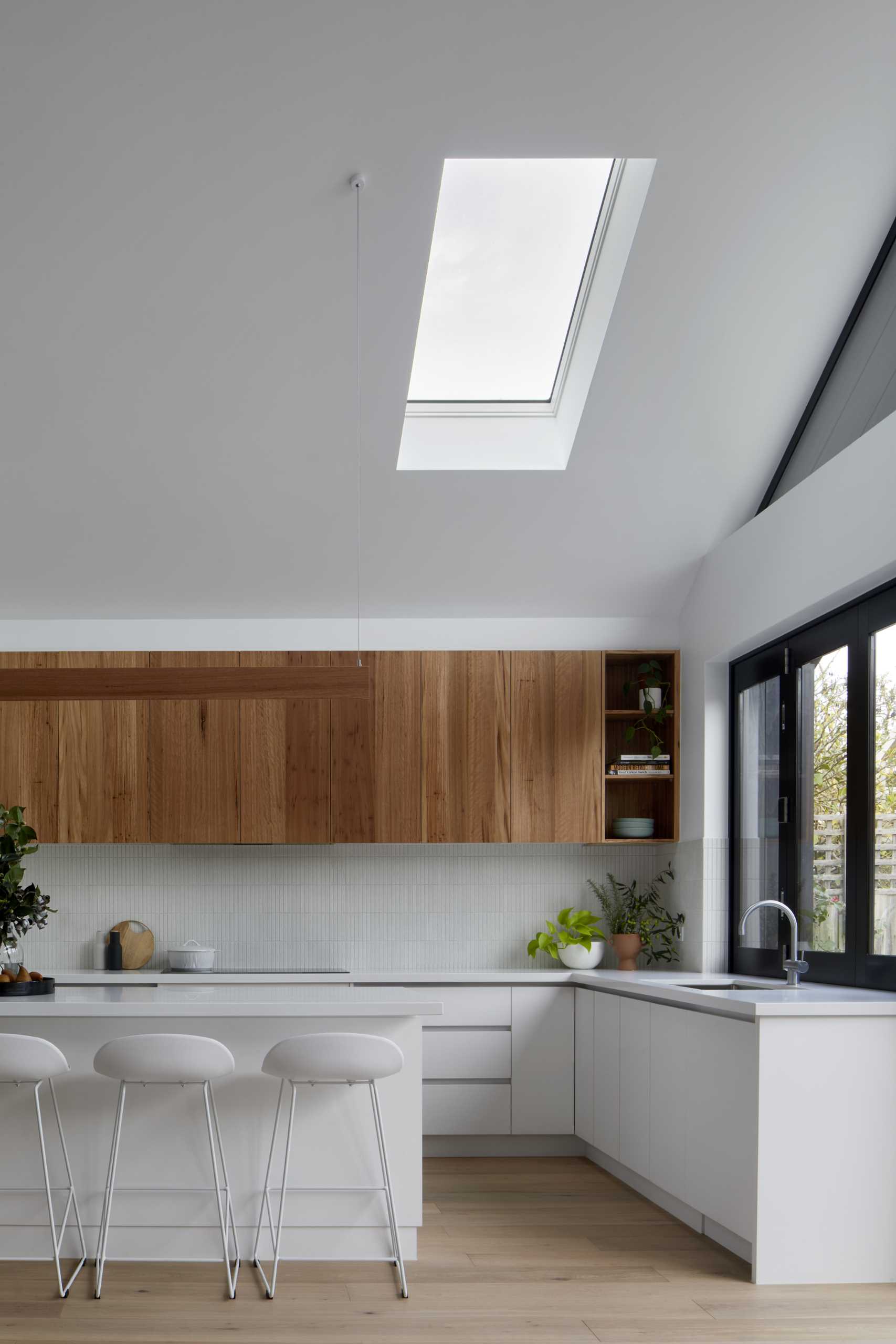 The folding pass-through window connects the kitchen to the outdoor space, while the rest of the kitchen includes white and wood cabinets, a skylight, and an island with open shelving.