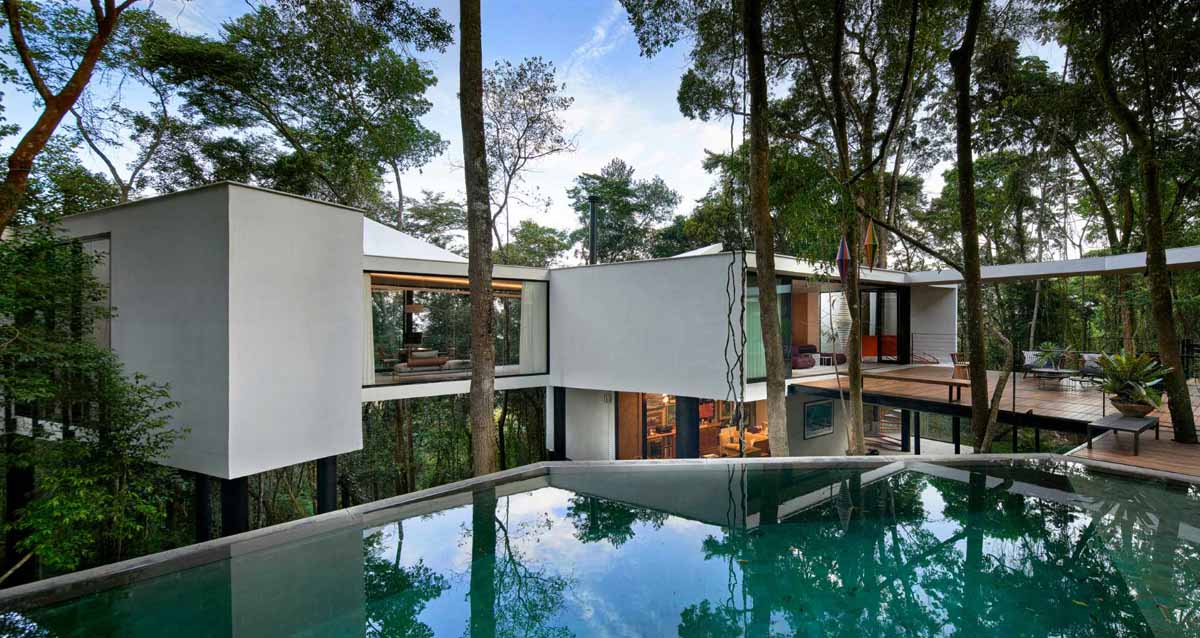 A modern house with a white exterior sits atop black stilts in the rainforest of Nova Lima, Brazil.