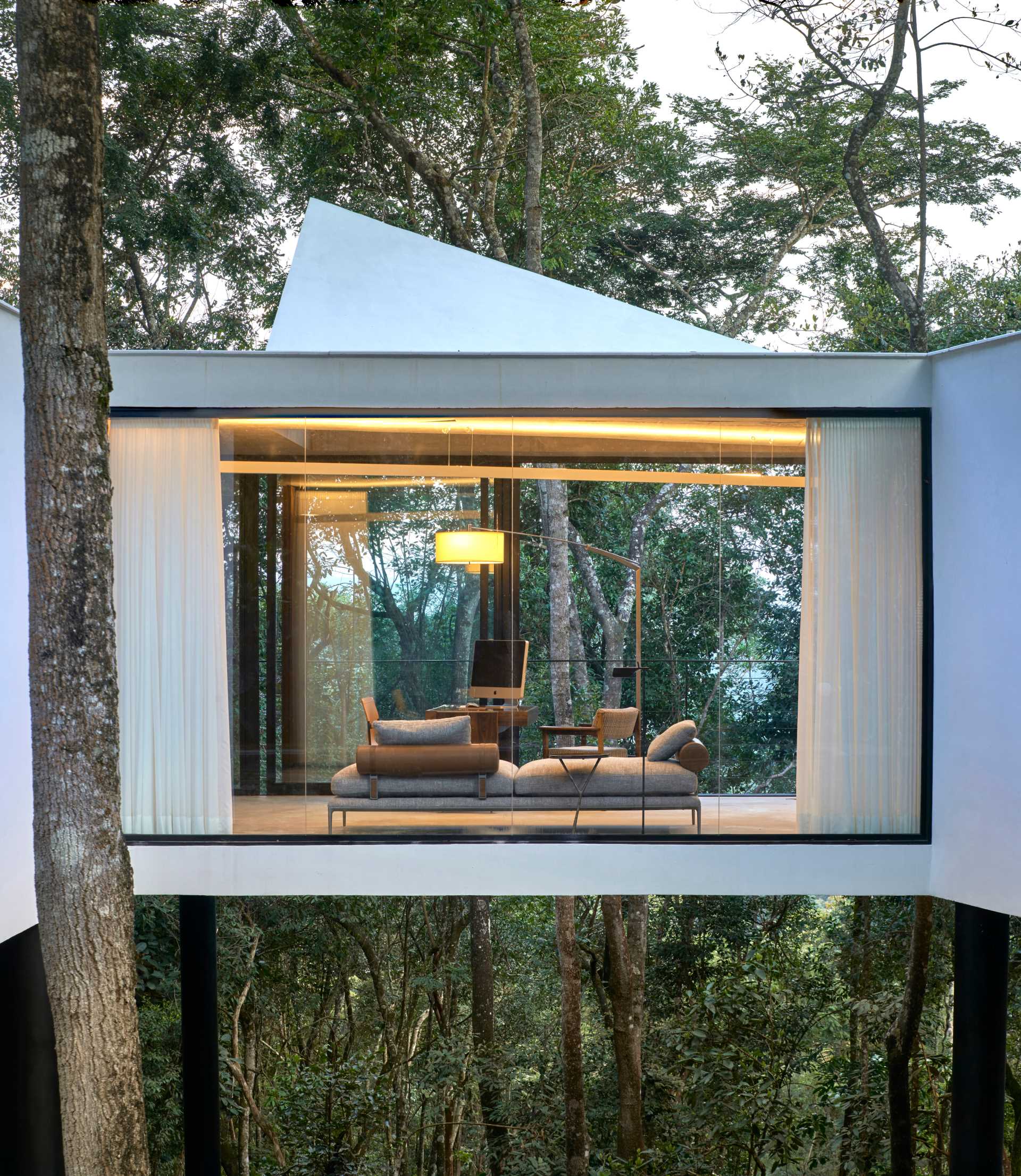 A modern house with a white exterior sits atop black stilts in the rainforest of Nova Lima, Brazil.