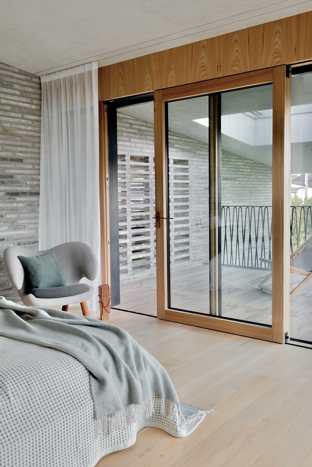 In this modern bedroom, the brick wall continues from the exterior through to the interior, while a balcony can be accessed through a sliding wood-framed glass door.