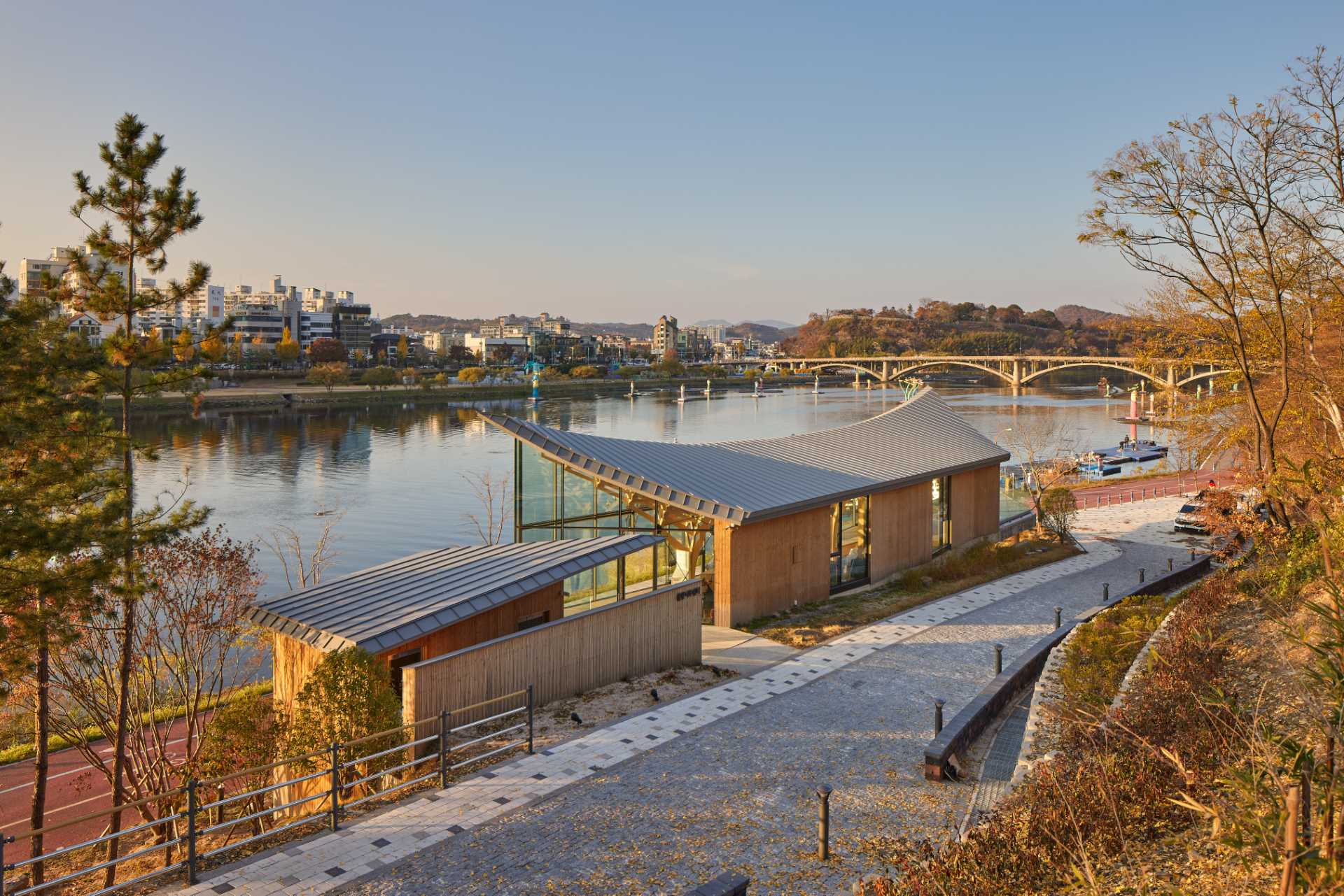 Six tree-like columns showcase wood craftsmanship inside a riverside pavilion with a glass front and curved roof.