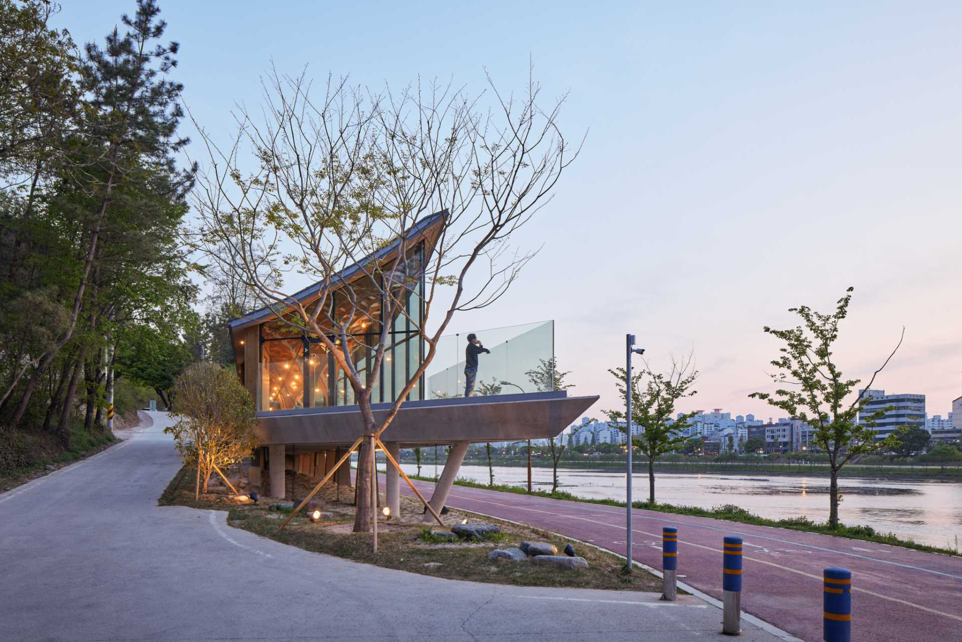 Six tree-like columns showcase wood craftsmanship inside a riverside pavilion with a glass front and curved roof.