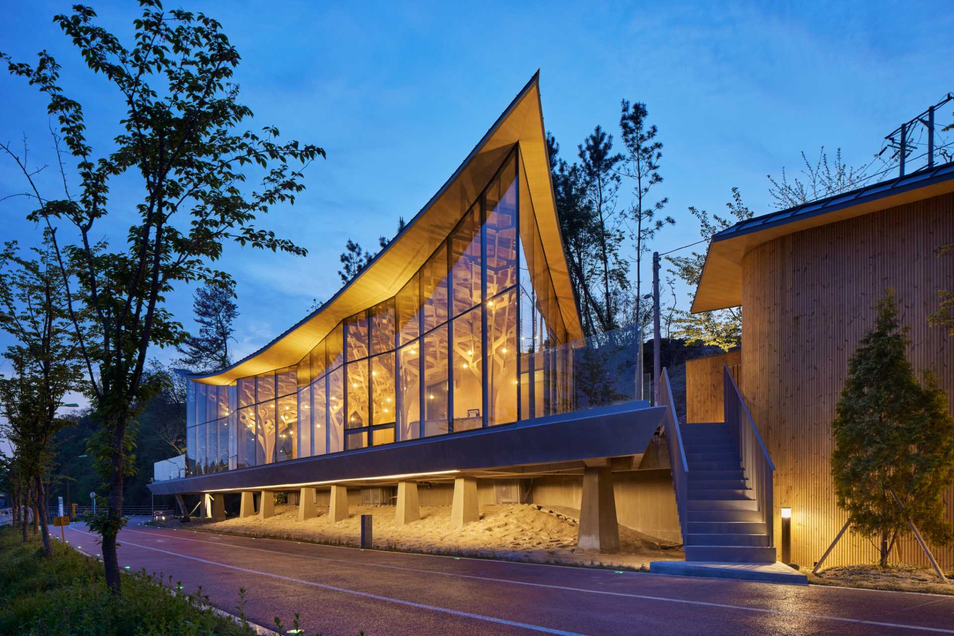 Six tree-like columns showcase wood craftsmanship inside a riverside pavilion with a glass front and curved roof.