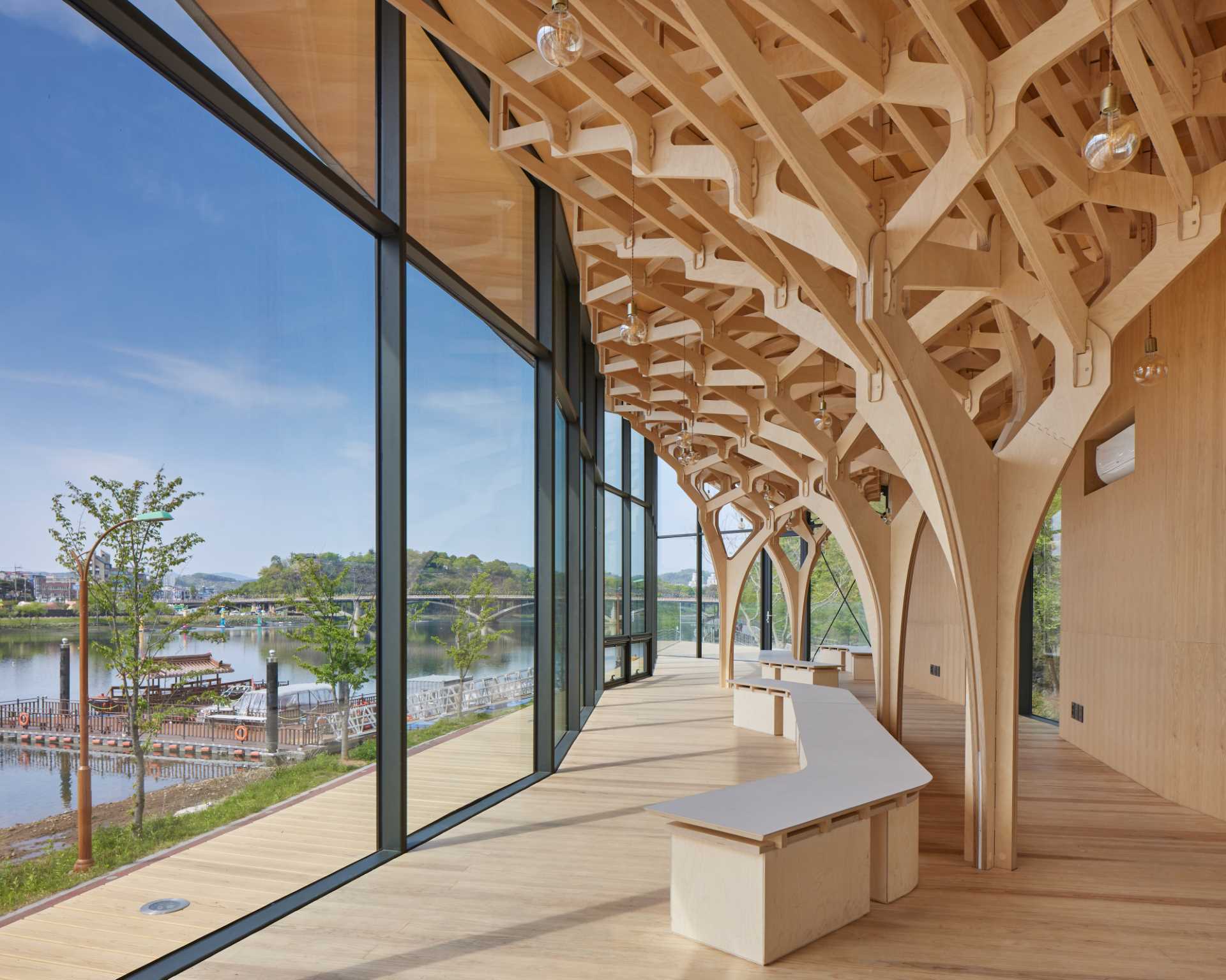 Six tree-like columns showcase wood craftsmanship inside a riverside pavilion with a glass front.