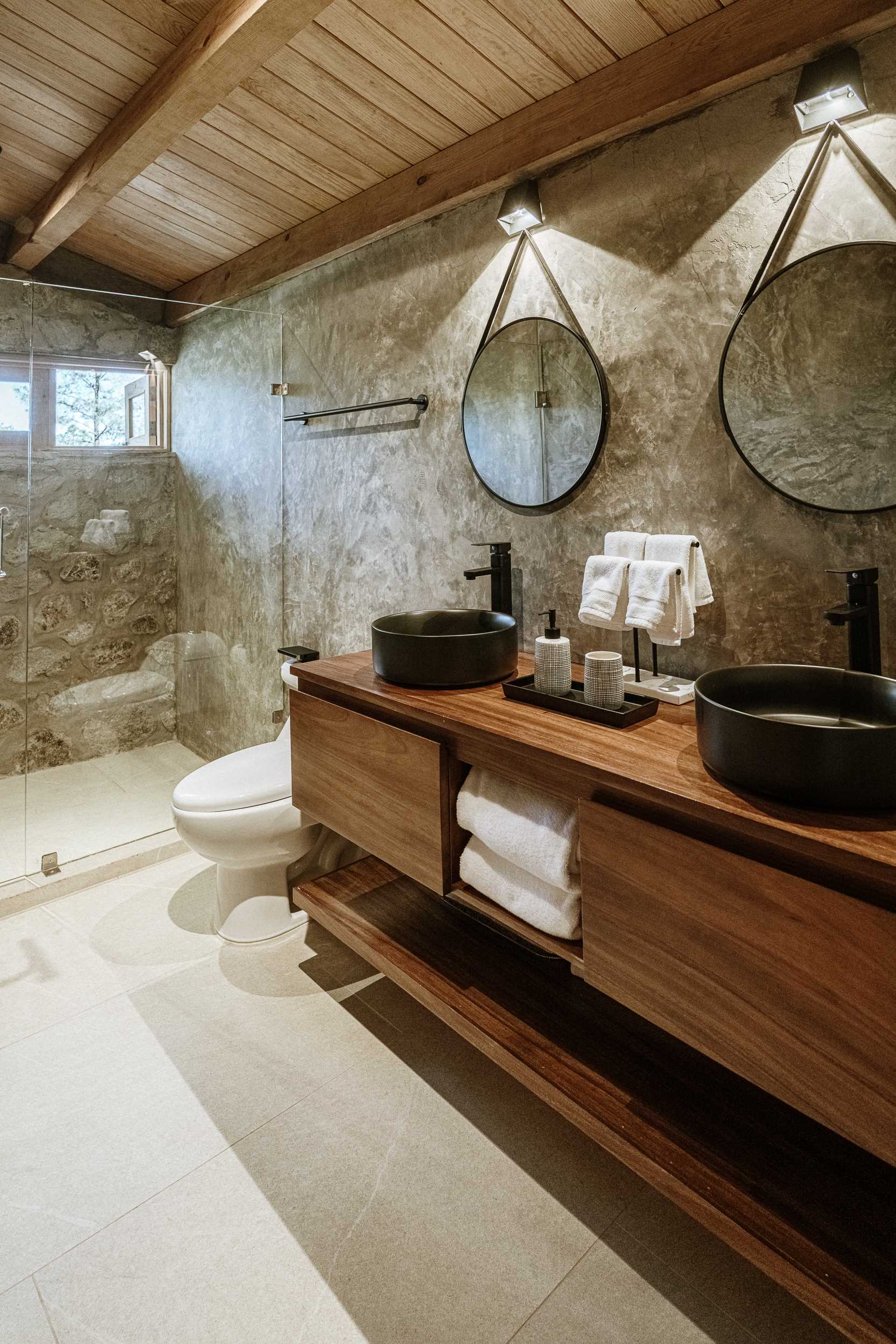 This stone and wood bathroom includes a double vanity and glass-enclosed shower.