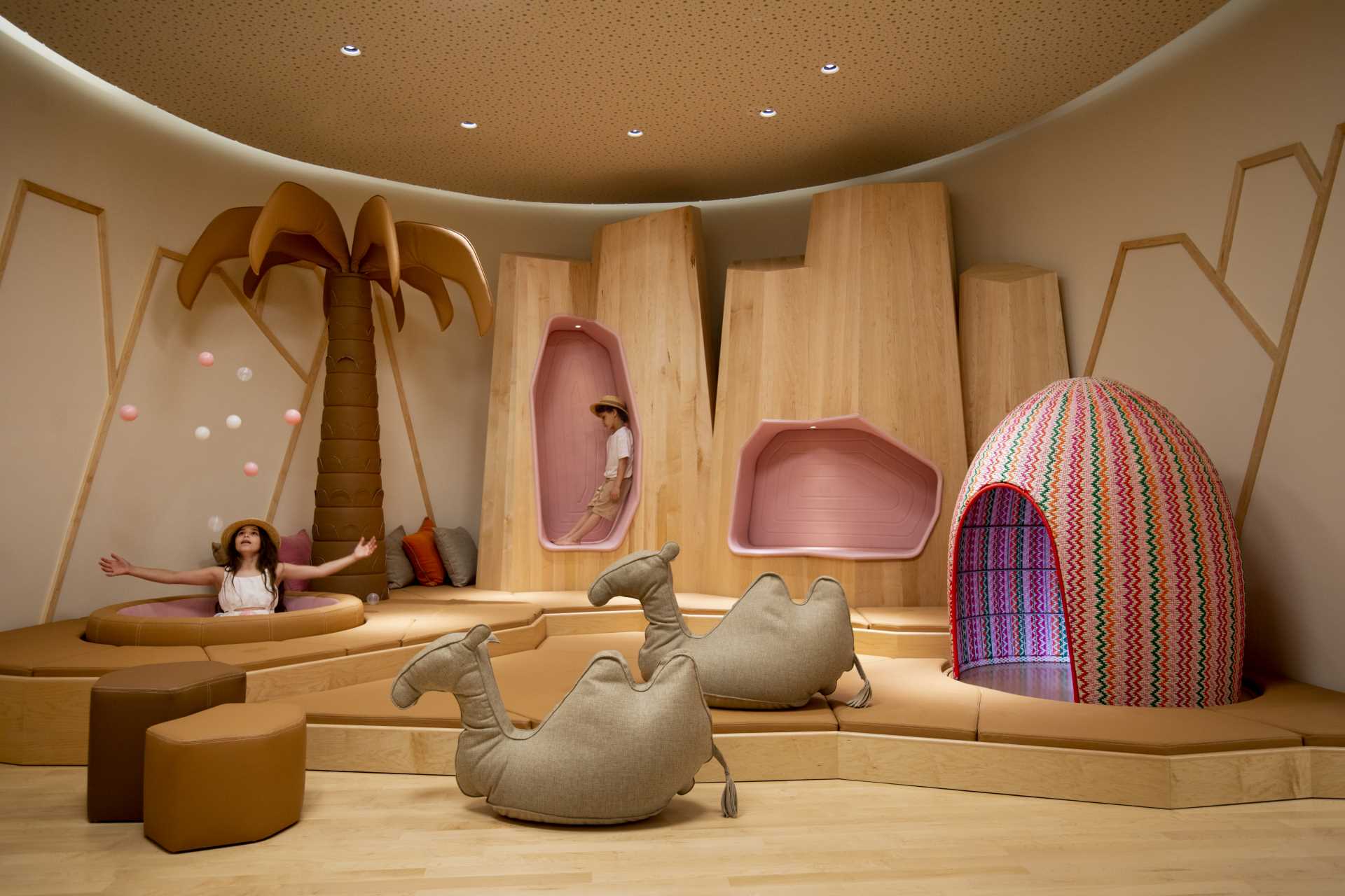 A modern kids playroom inspired by the desert, with sand dunes, sculptural desert mountains, camels, and palm trees.