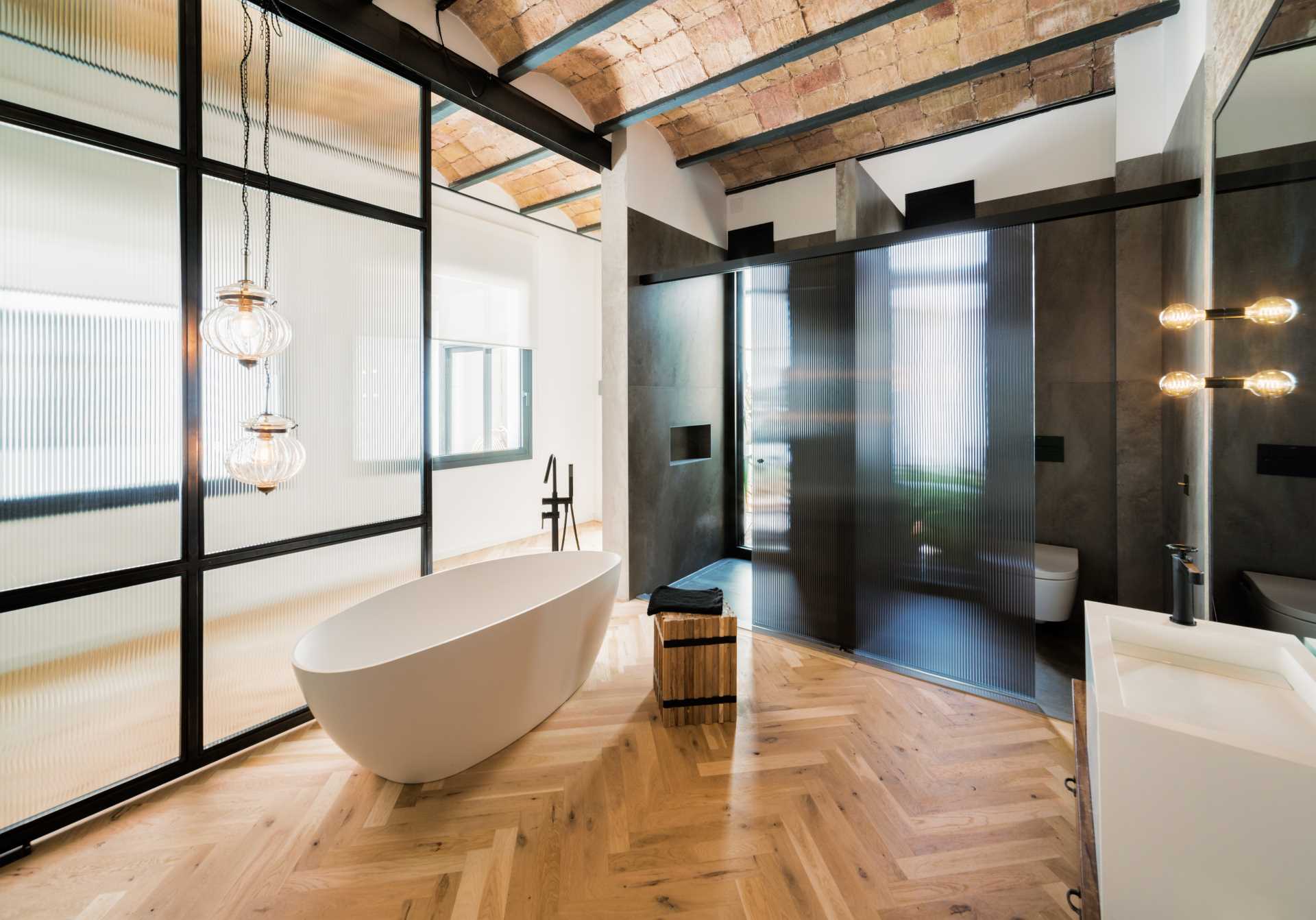 In this primary bedroom, herringbone flooring spans both the sleeping area and the bathroom, with a freestanding bathtub positioned in front of a large sliding glass door.