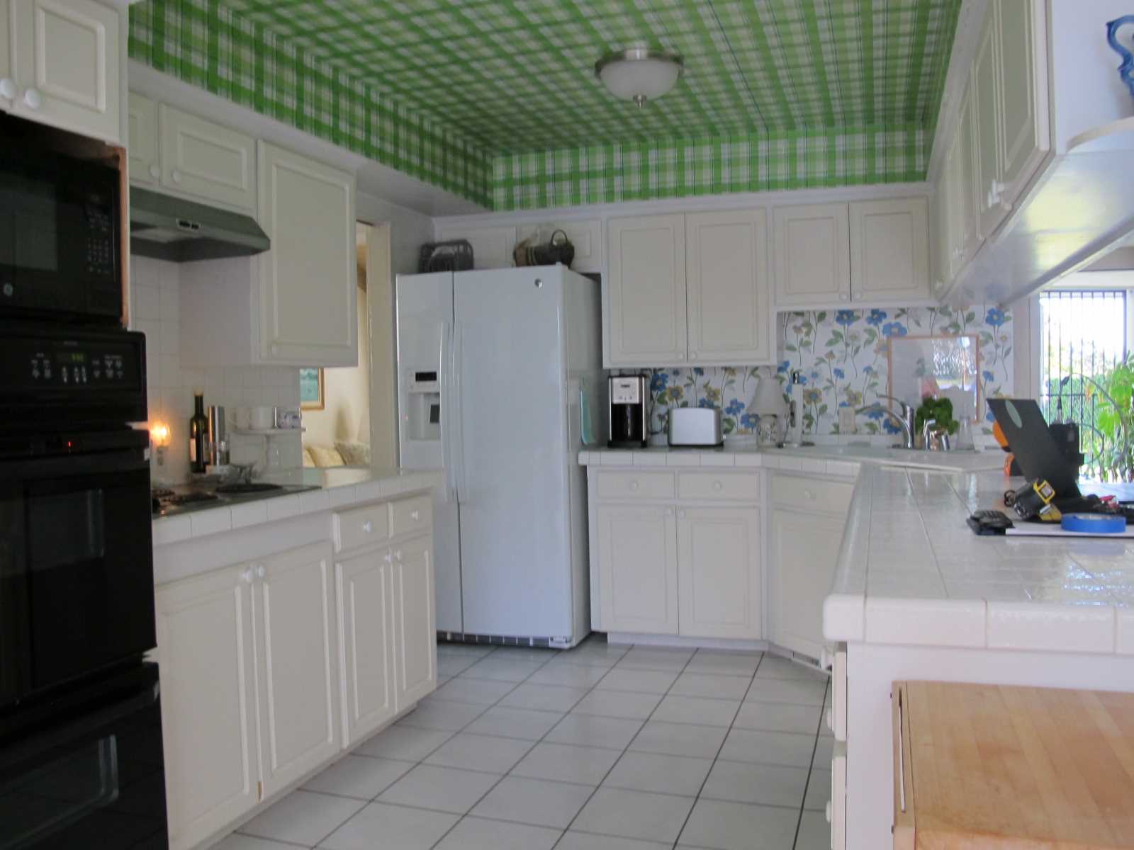 Before - The original kitchen had a tiled floor and countertops, with white cabinets, mismatched appliances, a green tartan wallpapered ceiling, and a floral wallpaper backsplash.