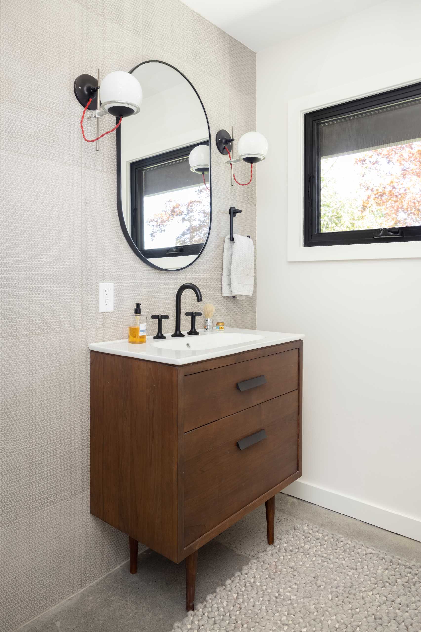In this updated bathroom, a tiled wall creates a backdrop for the dark wood vanity, while a black-framed oval mirror complements the window frame.