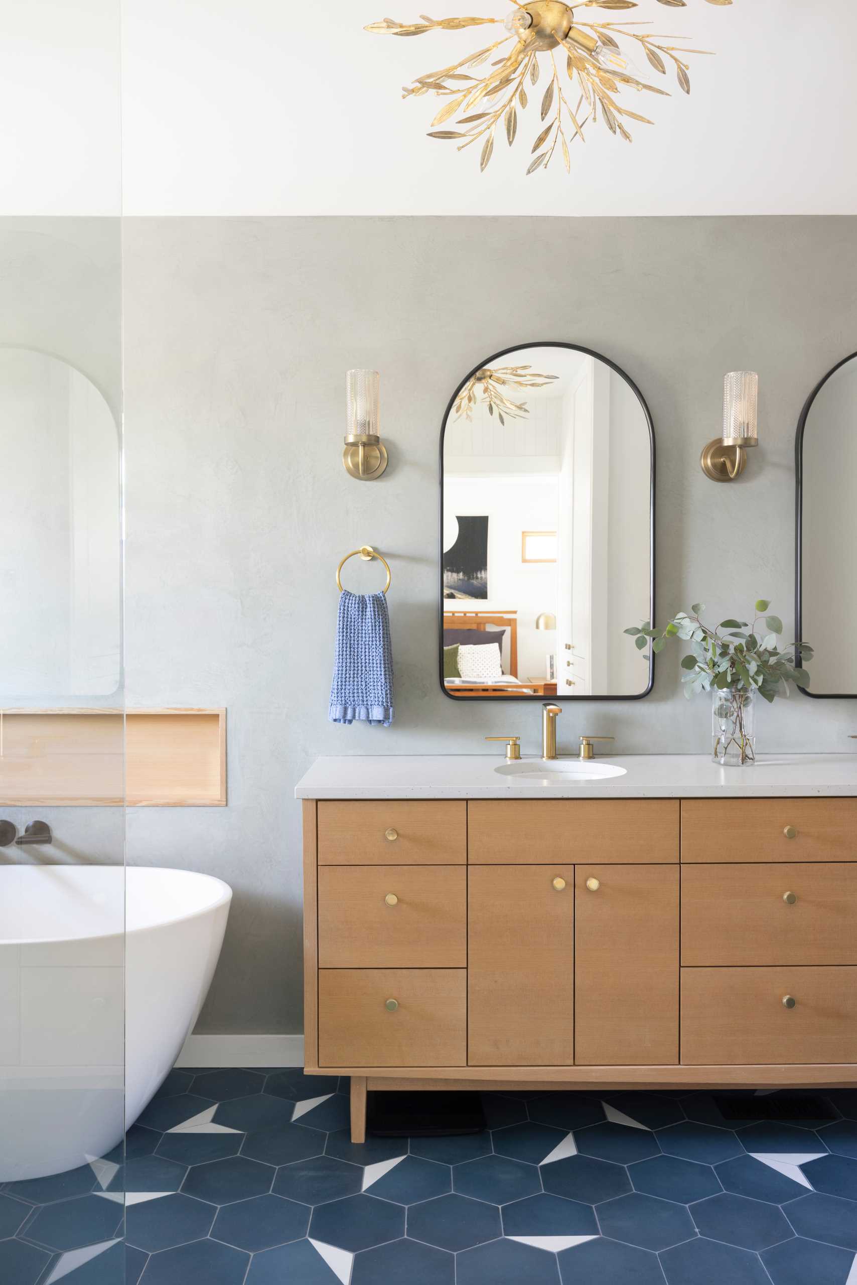 This ensuite bathroom includes a mid-century modern inspired tile flooring, a walk-in shower with a bench, white tiled walls, a freestanding bathtub with wood shelving niche, and a wood vanity.