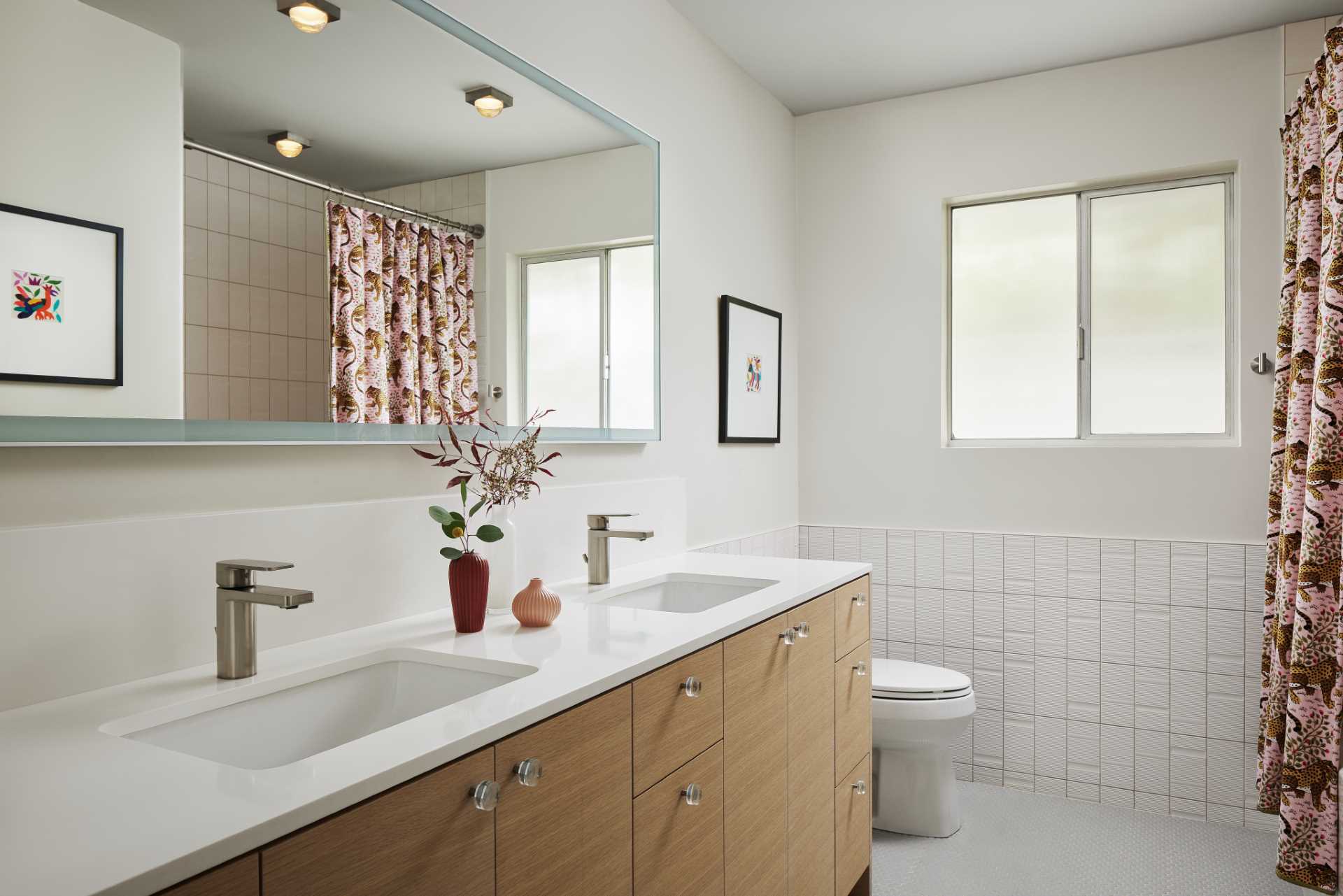 The updated bathroom is bright and open and now includes a double vanity with undermount sinks, a long horizontal mirror, and tile that wraps from the end of the vanity and around the wall to the shower.