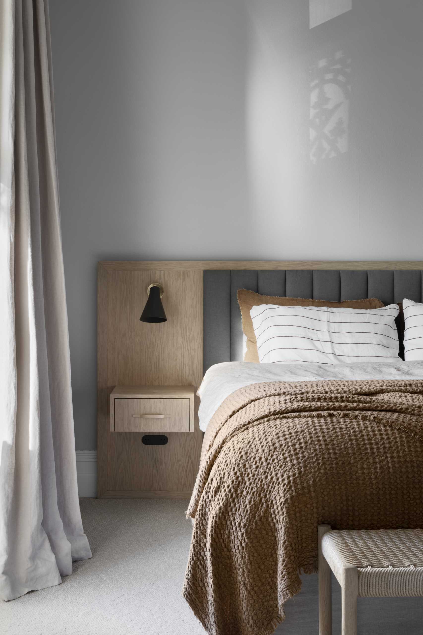 In an updated bedroom, a light wood and upholstered headboard includes a floating bedside table, as well as a wood closet and built-in desk.
