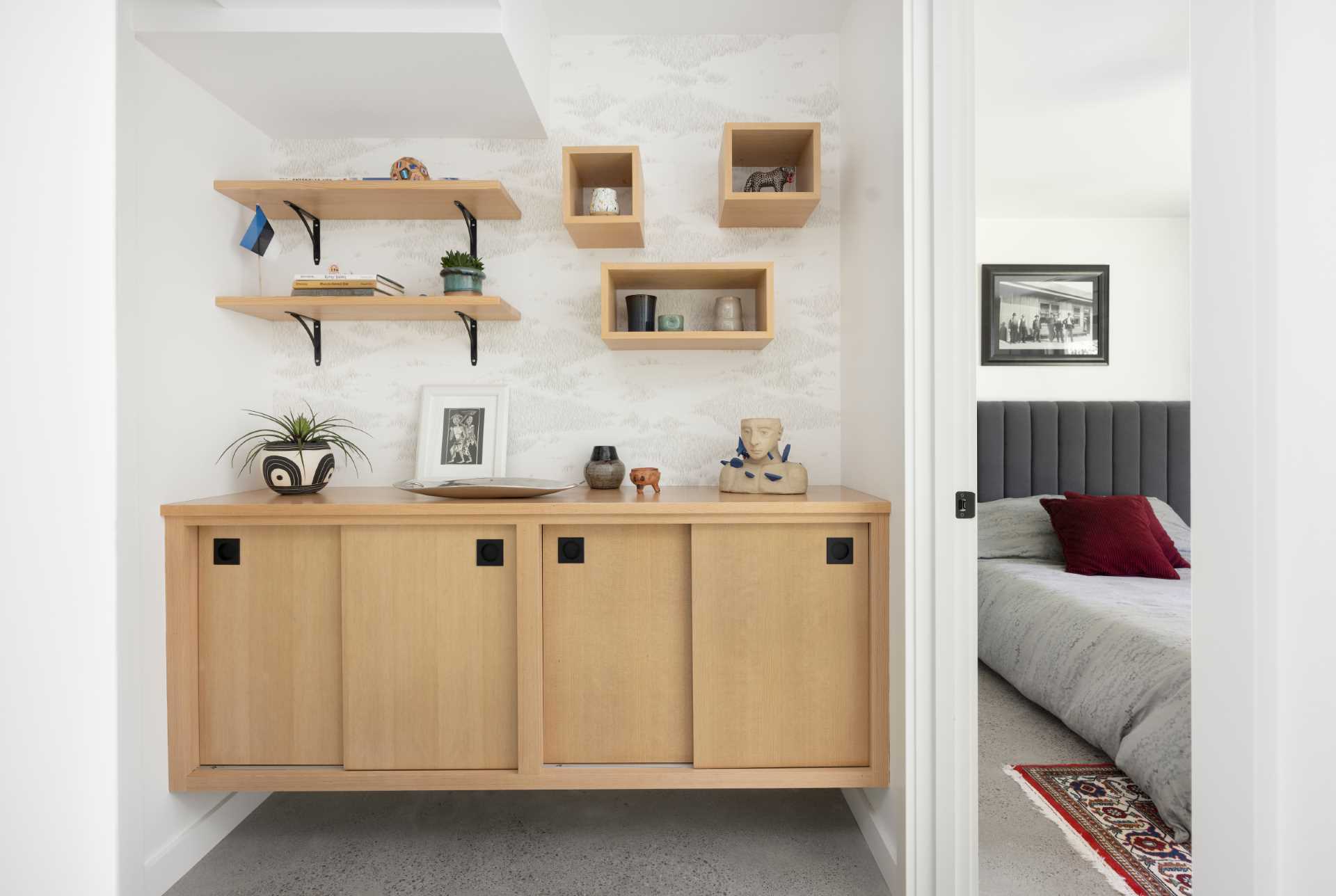 An updated interior includes a wood cabinet outside a bedroom, while inside the bedroom there's a dark grey upholstered headboard and a sliding door that opens to the outdoors.
