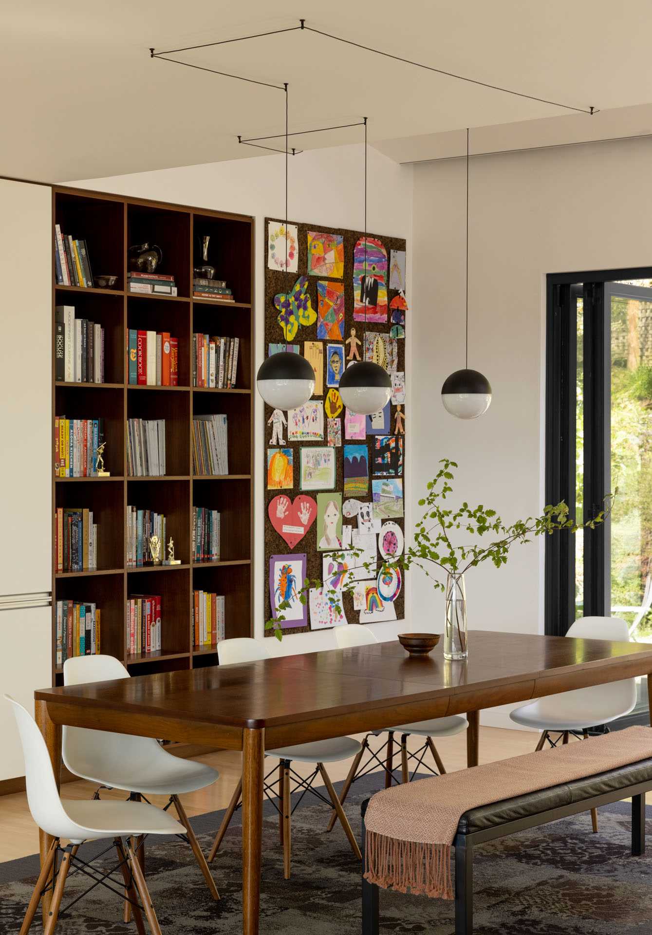 This modern dining room has a large wood dining table with bench and chair seating, as well as a wood bookshelf and art wall.