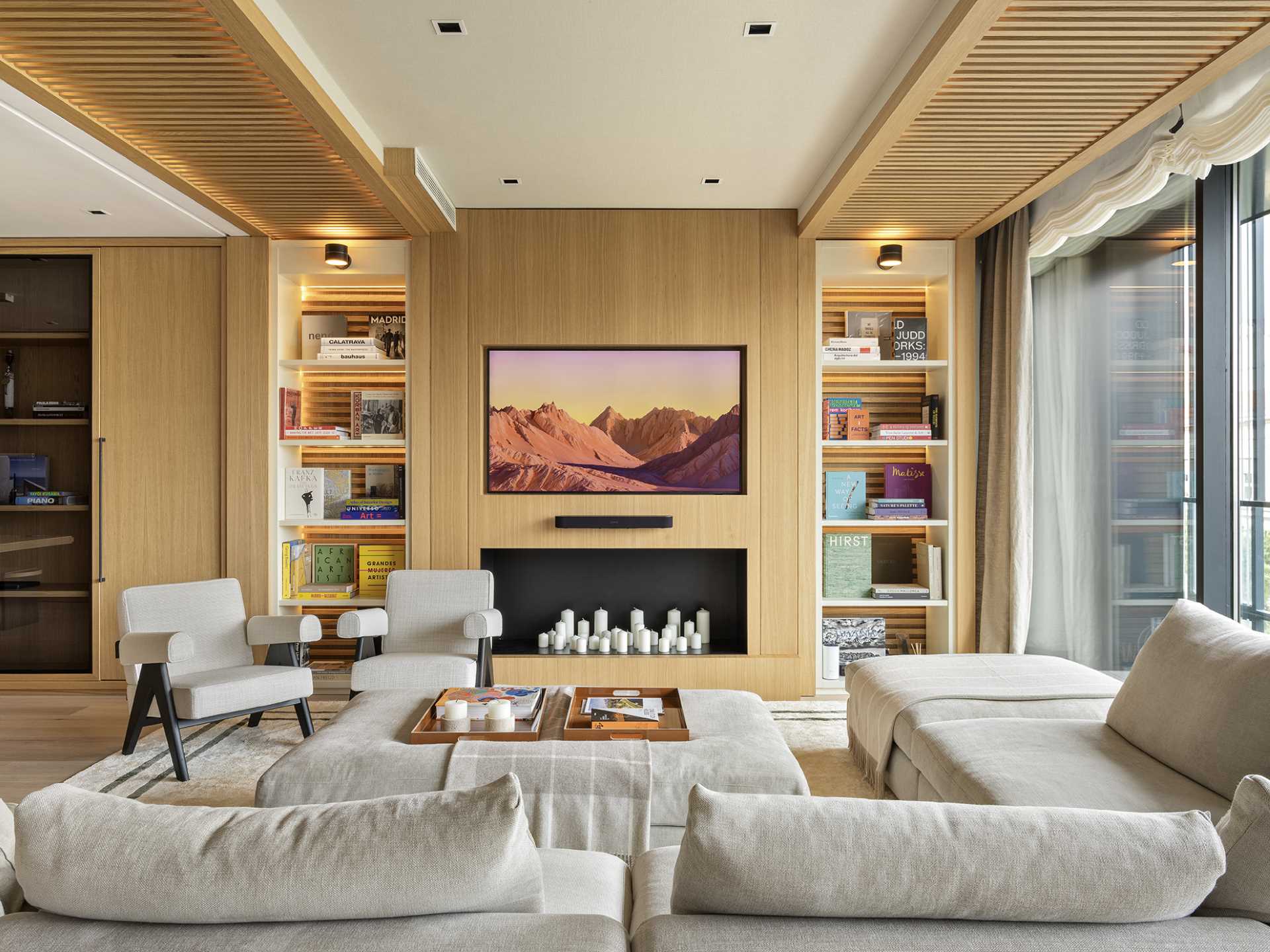 In the living room, a wall-mounted TV is disguised as art, while illuminated wooden shelves on either side stand as curated showcases, highlighting the owner's colorful book collection.