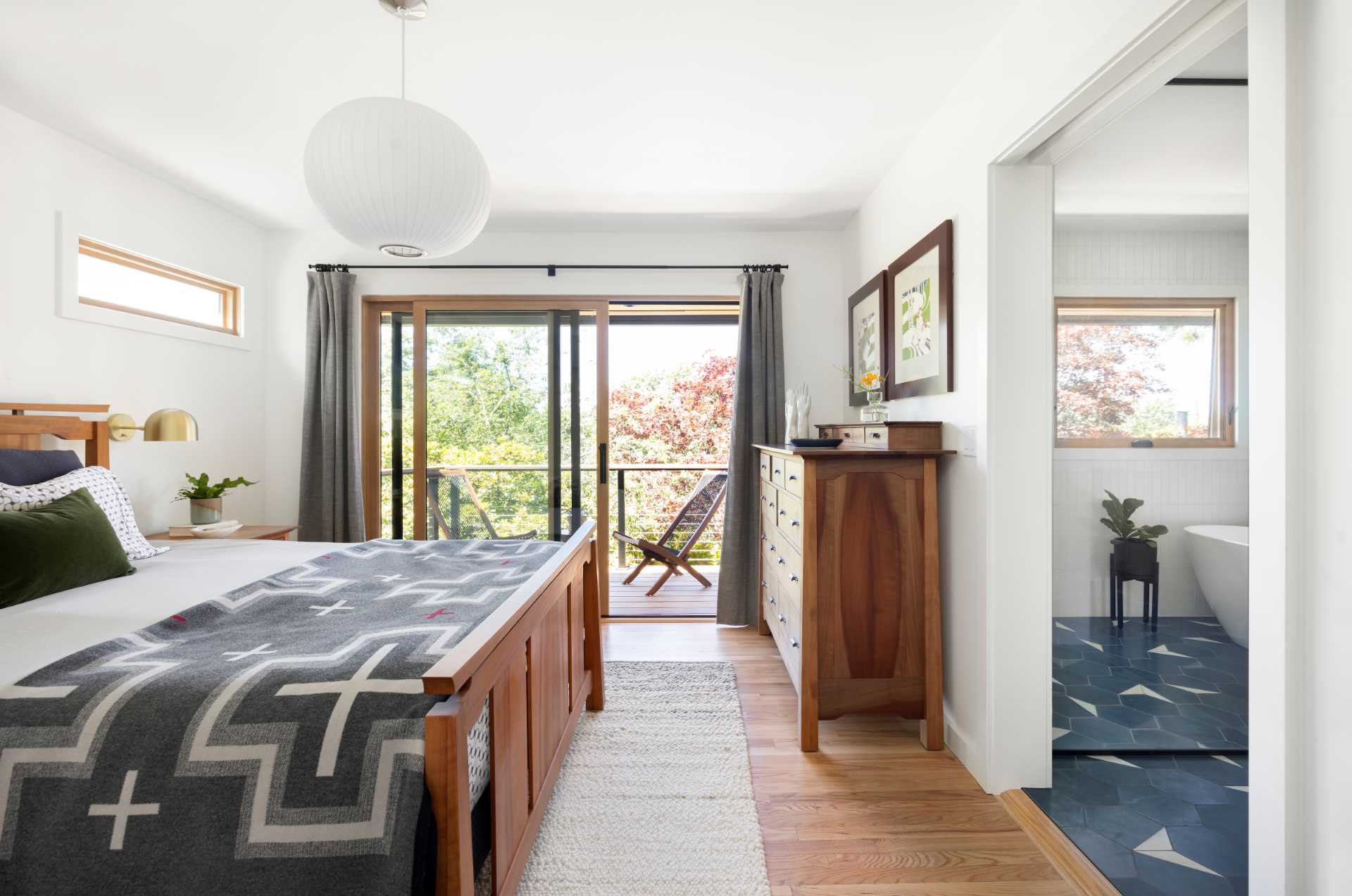 This updated primary bedroom has bright white walls and wood furniture that complements the wood window frames and door frame.