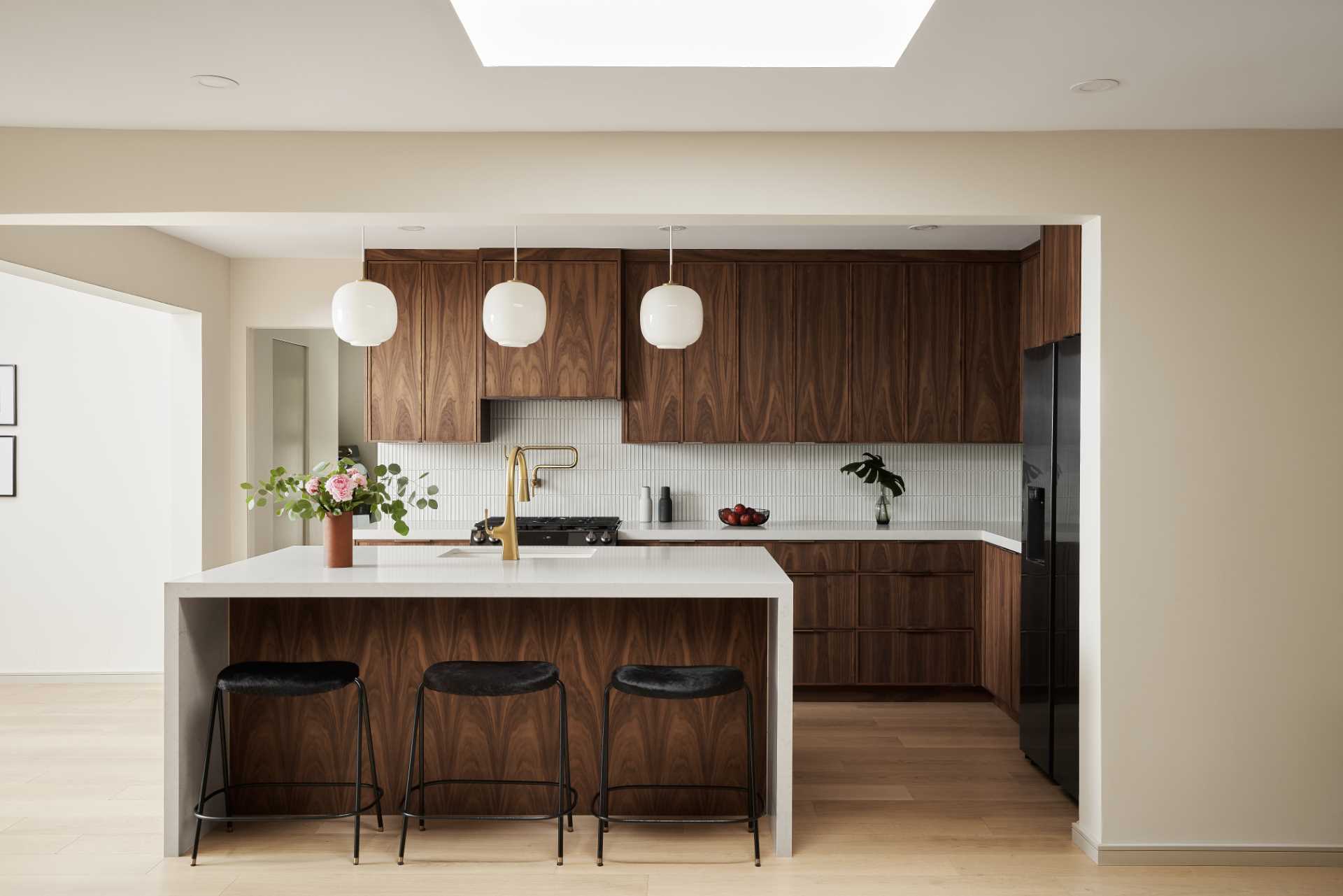 AFTER - This remodeled kitchen has been opened up to connect with the living room and dining room. New wood floors are featured throughout, and the warm custom walnut wood cabinets in the kitchen are paired with a thin vertical tile backsplash that wraps around the walls, while the countertop complements the large island.