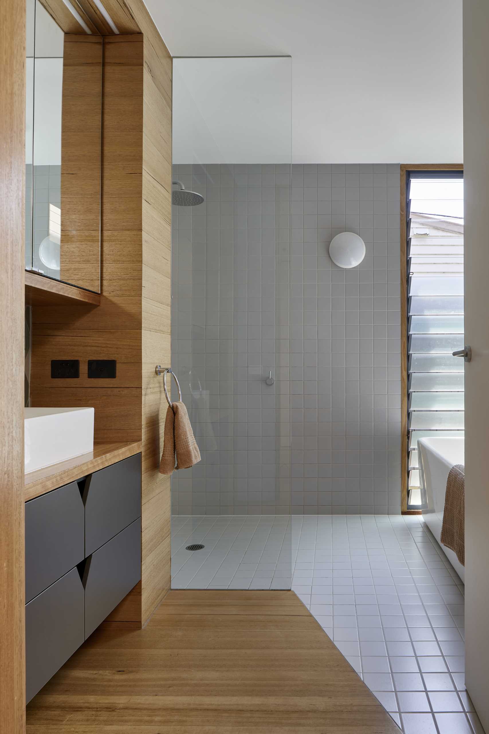 A modern bathroom includes a combination of wood flooring and square grey tiles that travel from the floor to the shower wall, while the vanity is built-in and includes dark grey cabinetry.