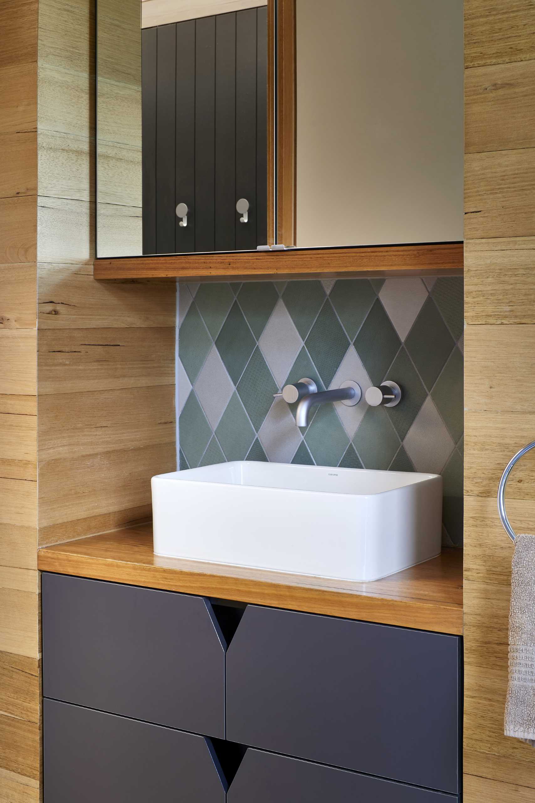 A modern bathroom includes a combination of wood flooring and square grey tiles that travel from the floor to the shower wall, while the vanity is built-in and includes dark grey cabinetry.