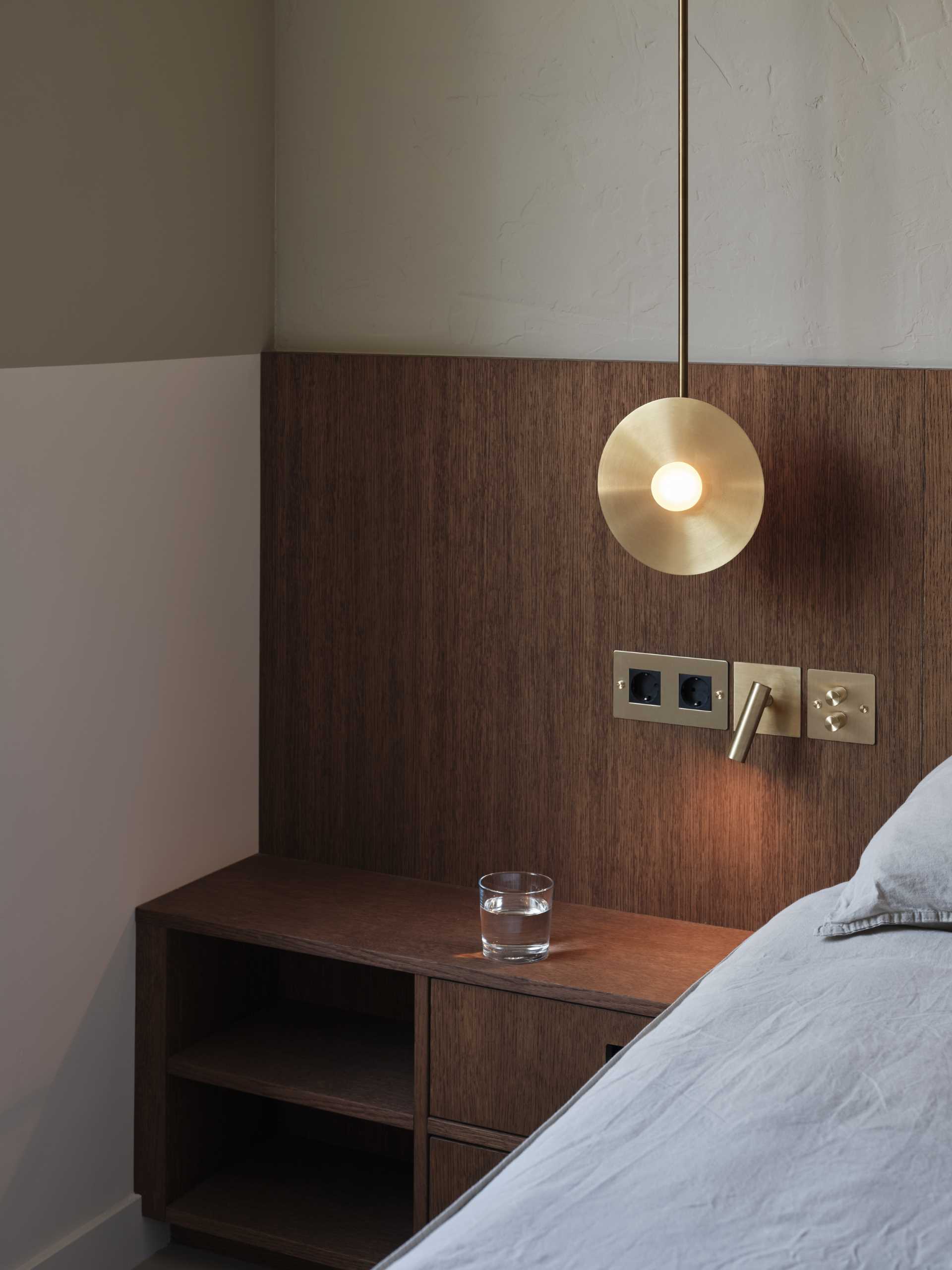 In this modern master bedroom, there are custom wooden cabinets, a tall mirror, rustic plaster, and a wooden bed frame. All of which are complimented by playful bedside lamps on each side of the bed.