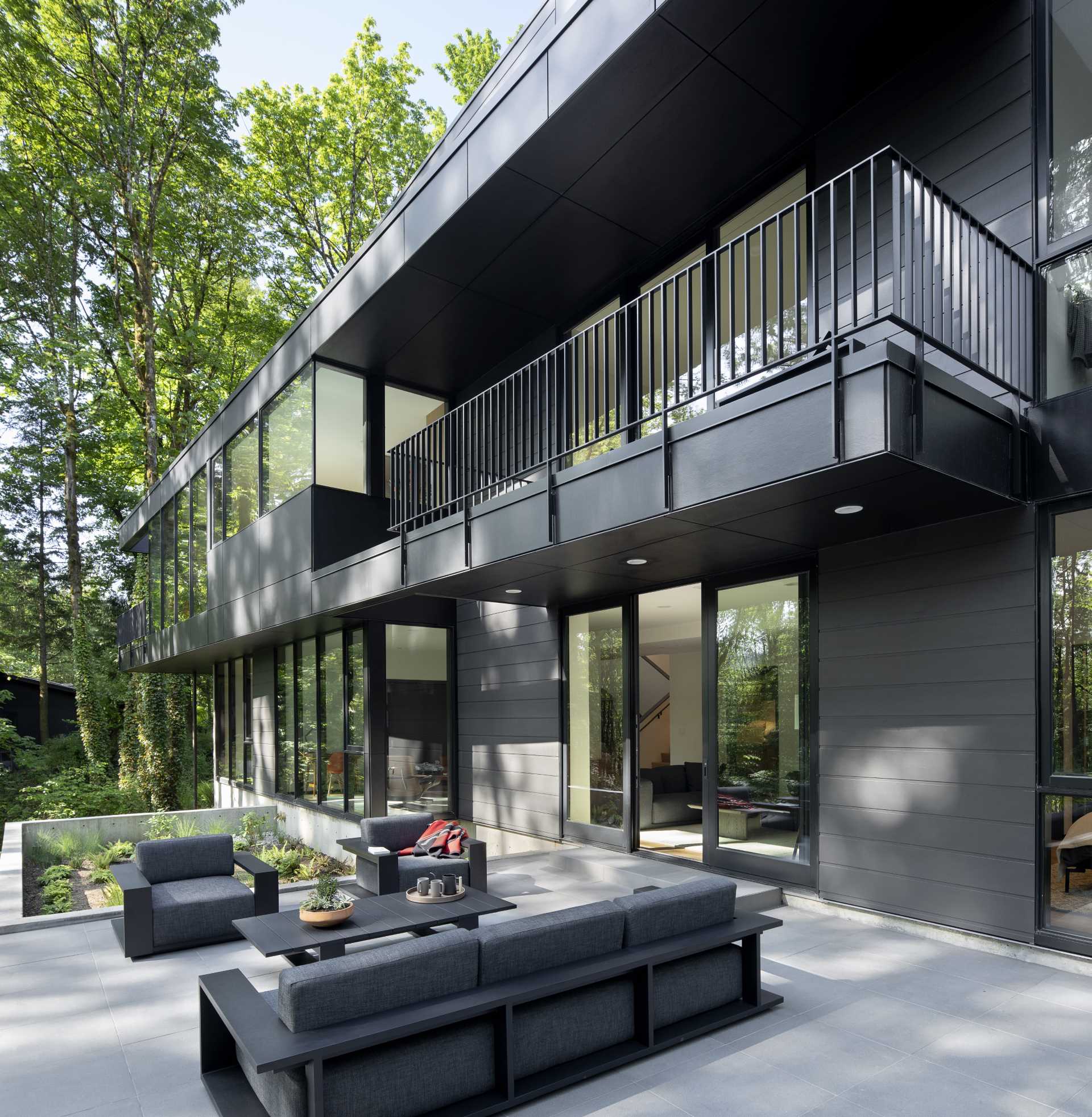 A modern black house with rooms that open to a patio furnished with outdoor furniture, and the forest beyond.