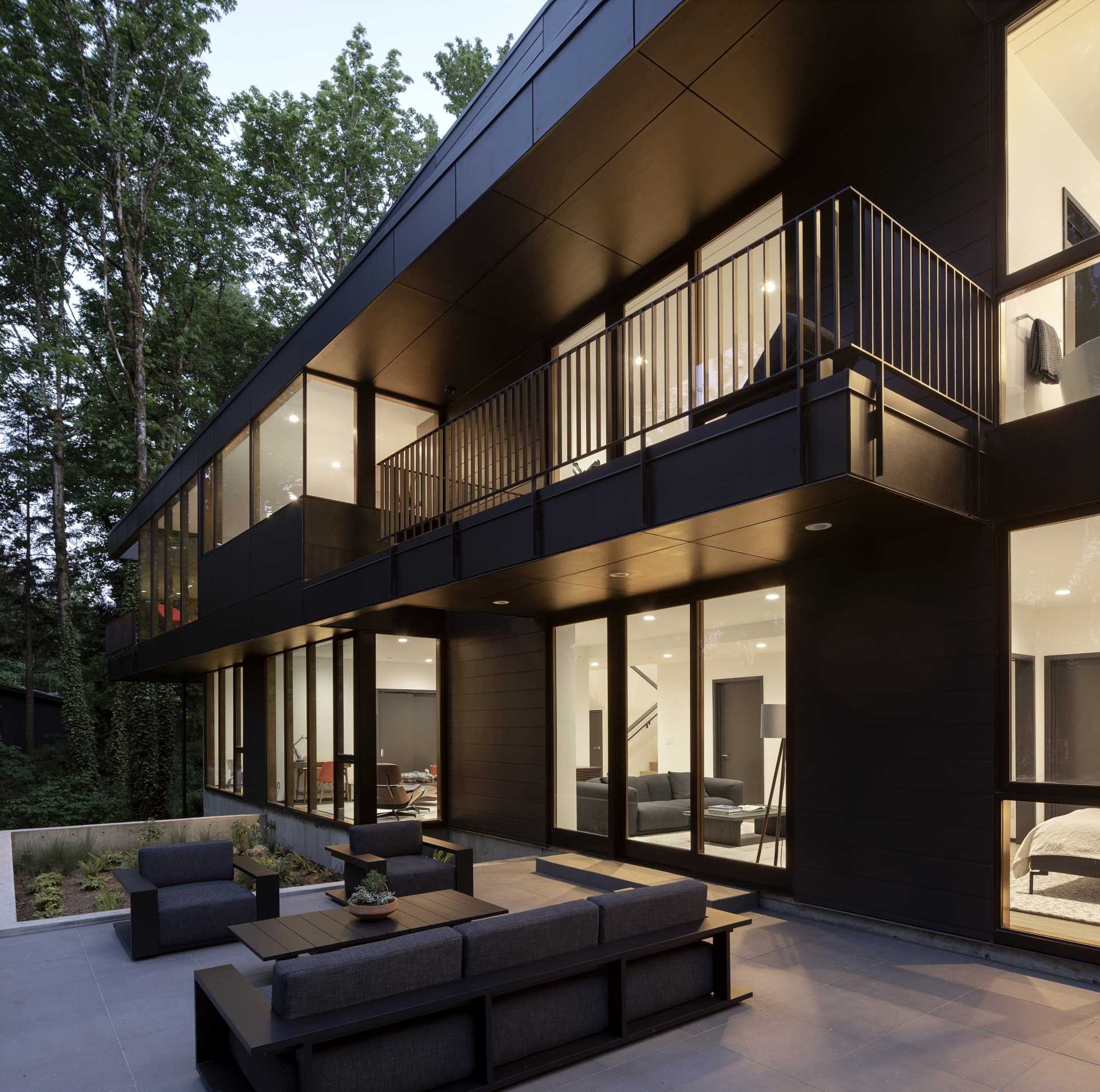 A modern black house with rooms that open to a patio furnished with outdoor furniture, and the forest beyond.