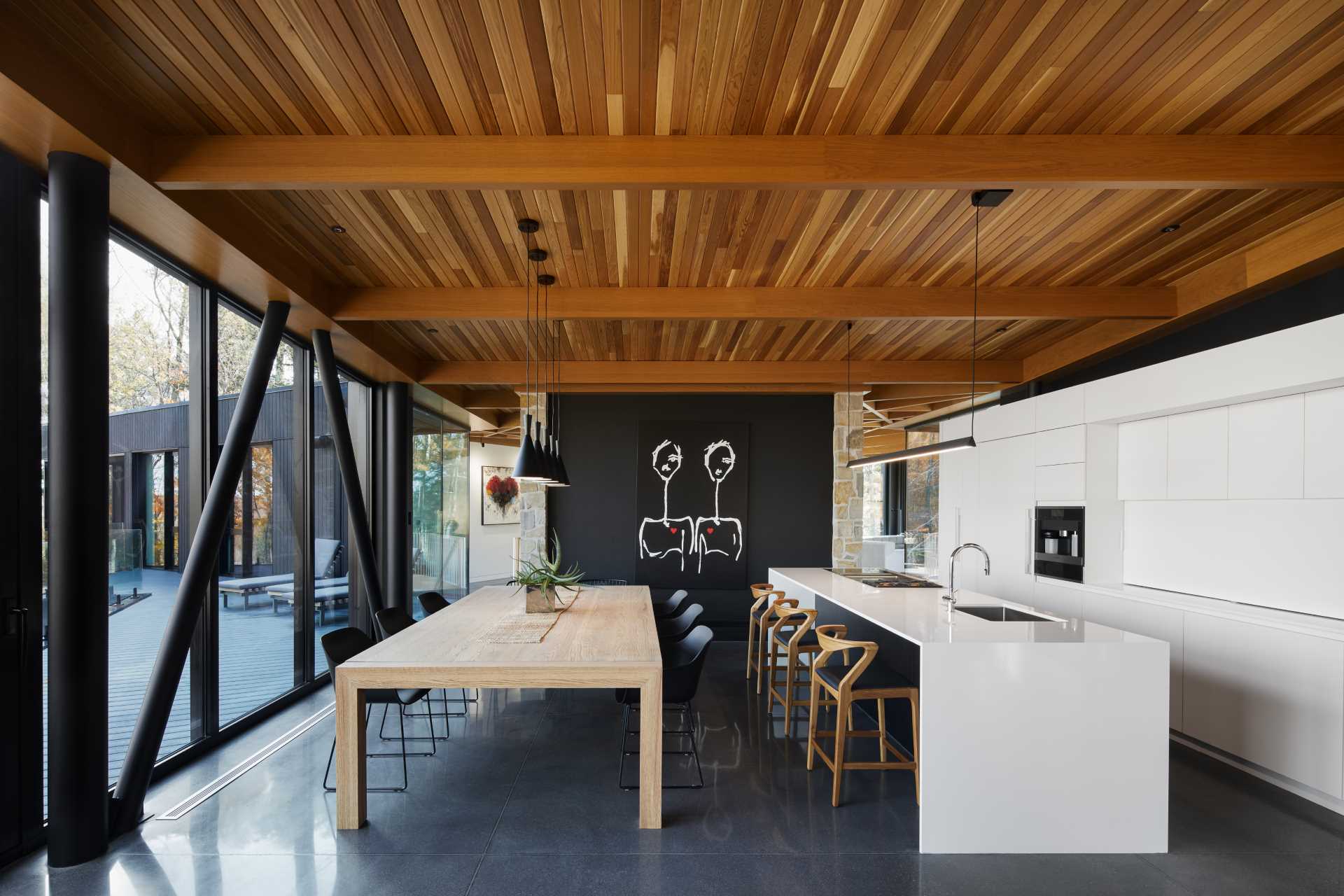 The light materials of this open-plan dining area and kitchen contrast the black accents of the structure, walls, and exterior.
