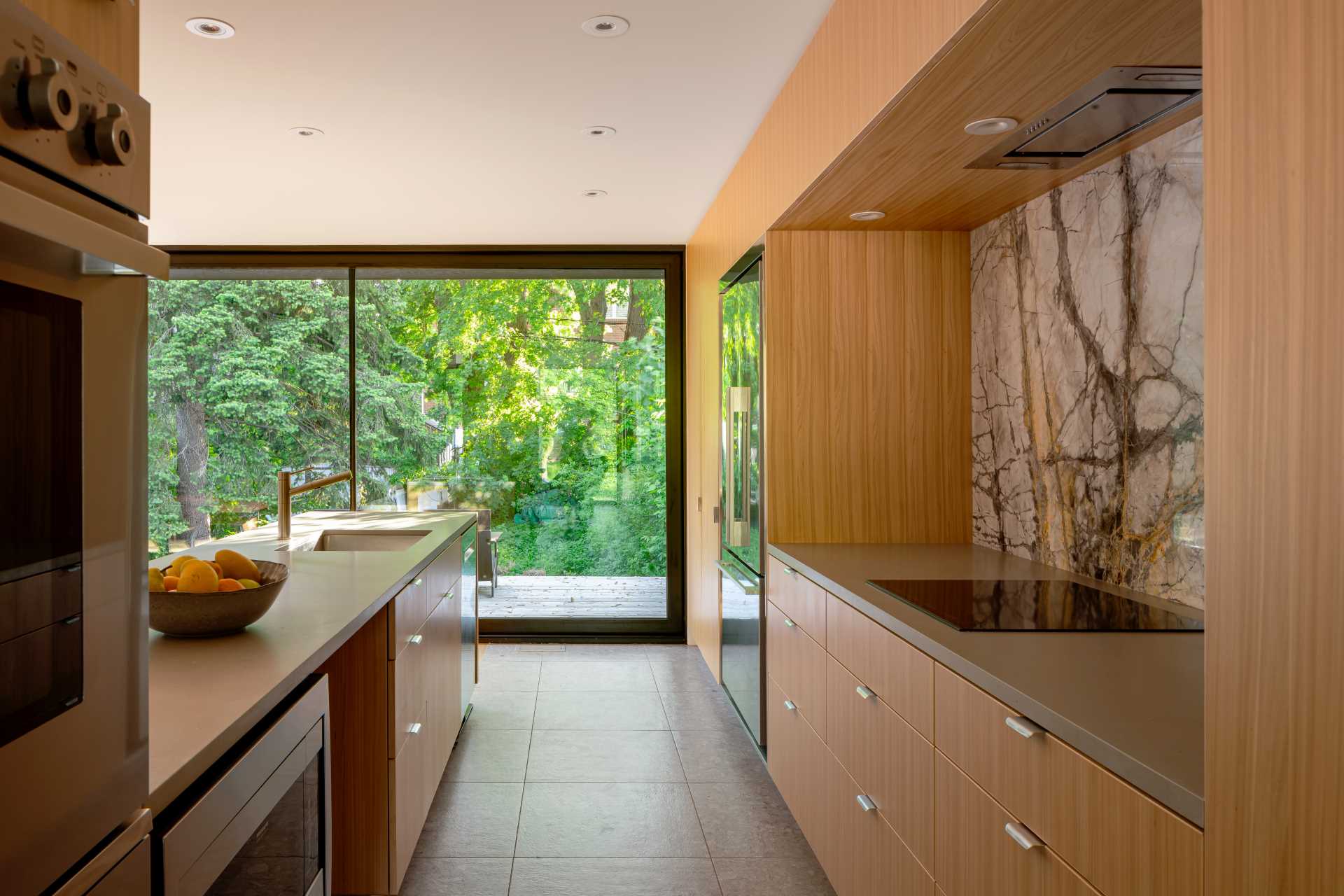 In this kitchen with wood cabinets, large floor-to-ceiling openings provide natural light and views of the green garden areas beyond.