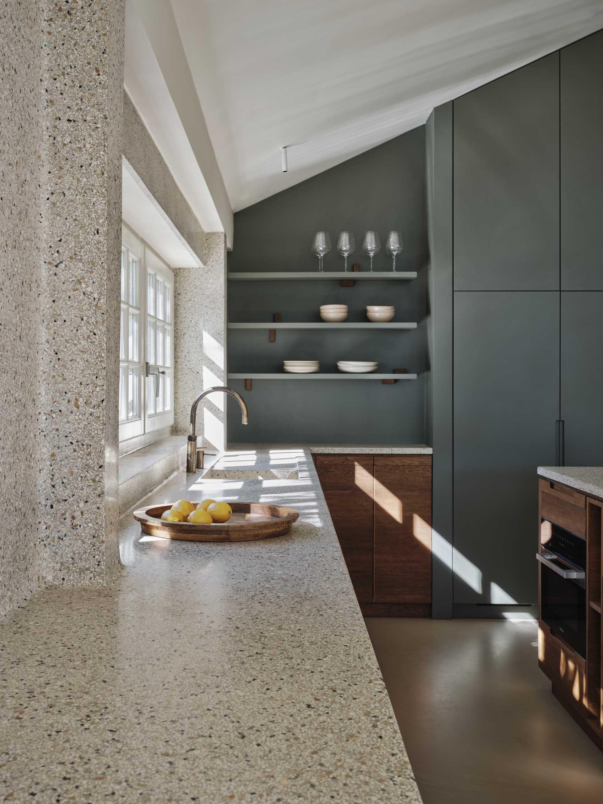 A modern kitchen with dark wood and matte green cabinets, also includes a seamless terrazzo countertop and integrated sink, that then travels up onto the wall.