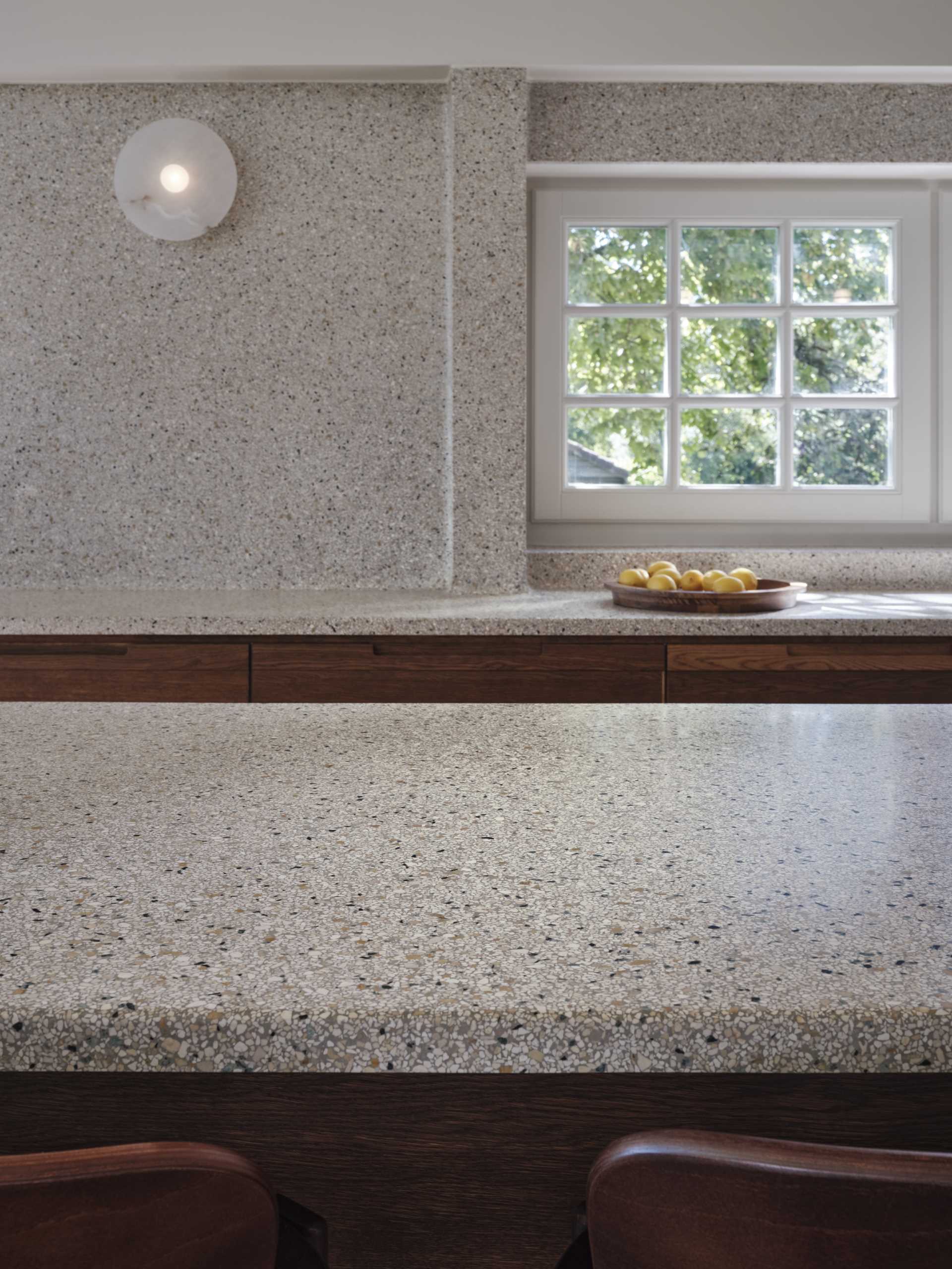 A modern kitchen with dark wood and matte green cabinets, also includes a seamless terrazzo countertop and integrated sink, that then travels up onto the wall.